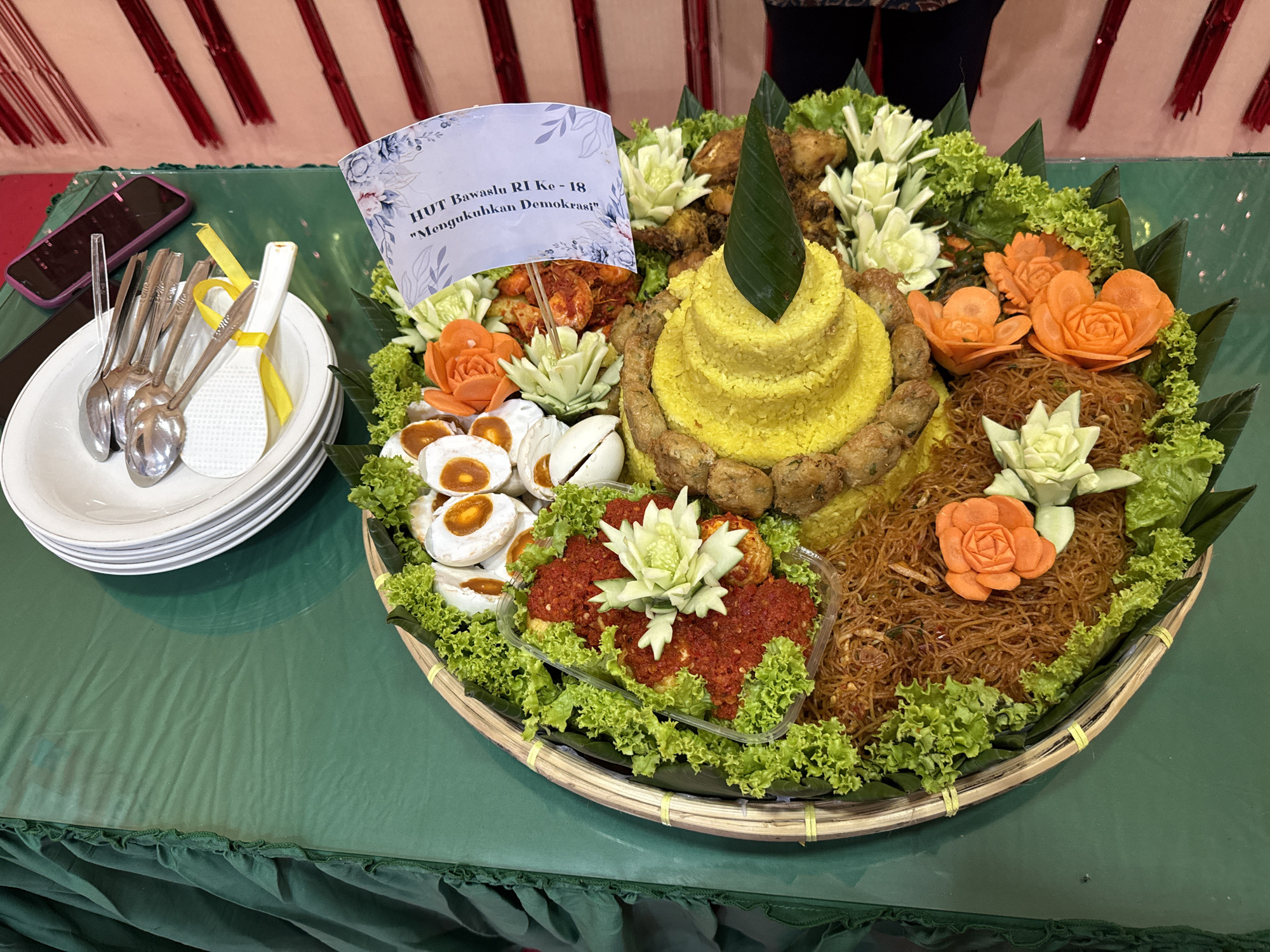 The image depicts a Tumpeng dish, a traditional Indonesian cone-shaped rice dish, displayed on a woven tray, on top of a green tablecloth. The Tumpeng is adorned with various side dishes and vegetables, including fried chicken, noodles, and hard-boiled eggs. A small flag with Indonesian text is placed on the Tumpeng. Next to the Tumpeng is a stack of white plates with silverware. A phone is adjacent to the plates. The scene is set indoors. Given that the text on the flag includes "Bawaslu RI," referring to the Indonesian Election Supervisory Body, and based on the food culture, the image is likely from Indonesia. There is no information within the image to confirm or deny this image was taken in Lhokseumawe.