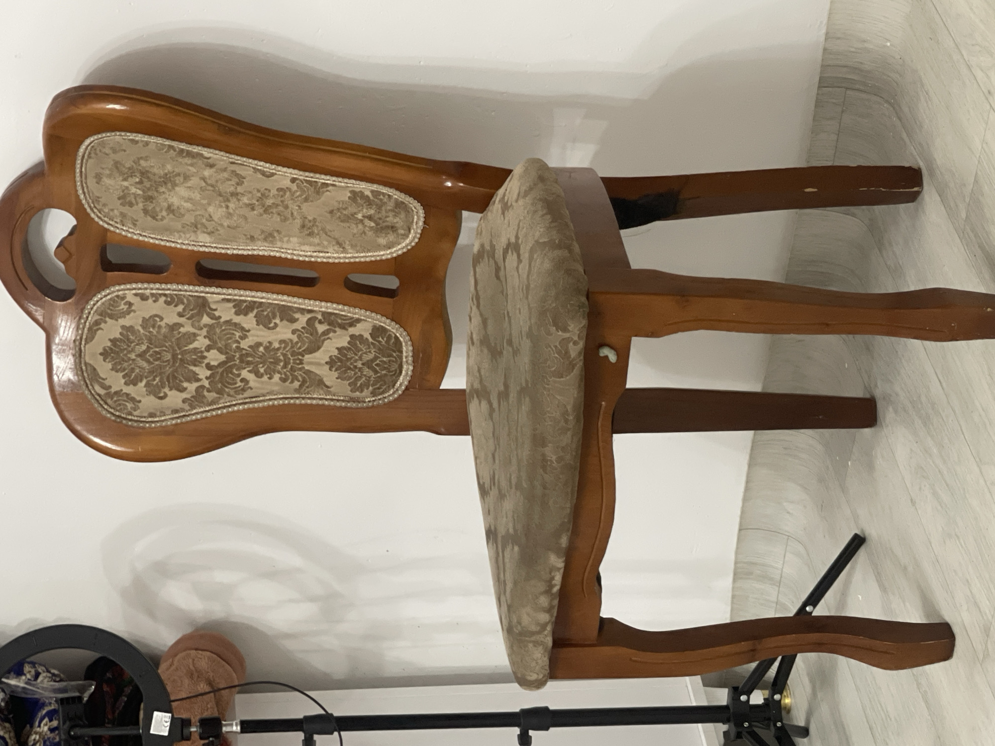 A single, ornate wooden chair with upholstered seat and back is positioned in front of a plain white wall. The chair has a light brown wood finish and features decorative fabric with a floral pattern on both the seat and the back. The backrest is high and has a carved handle-like opening at the top. The chair is illuminated from the left, casting a subtle shadow on the wall behind it. In the background, to the left of the chair, a black tripod with a ring light is visible, suggesting the setting might be a studio or an area prepared for photography or videography. The floor is a light-colored laminate with a wood grain pattern. No people or animals are present in the image. The location is described as Bishkek City, Kyrgyzstan.