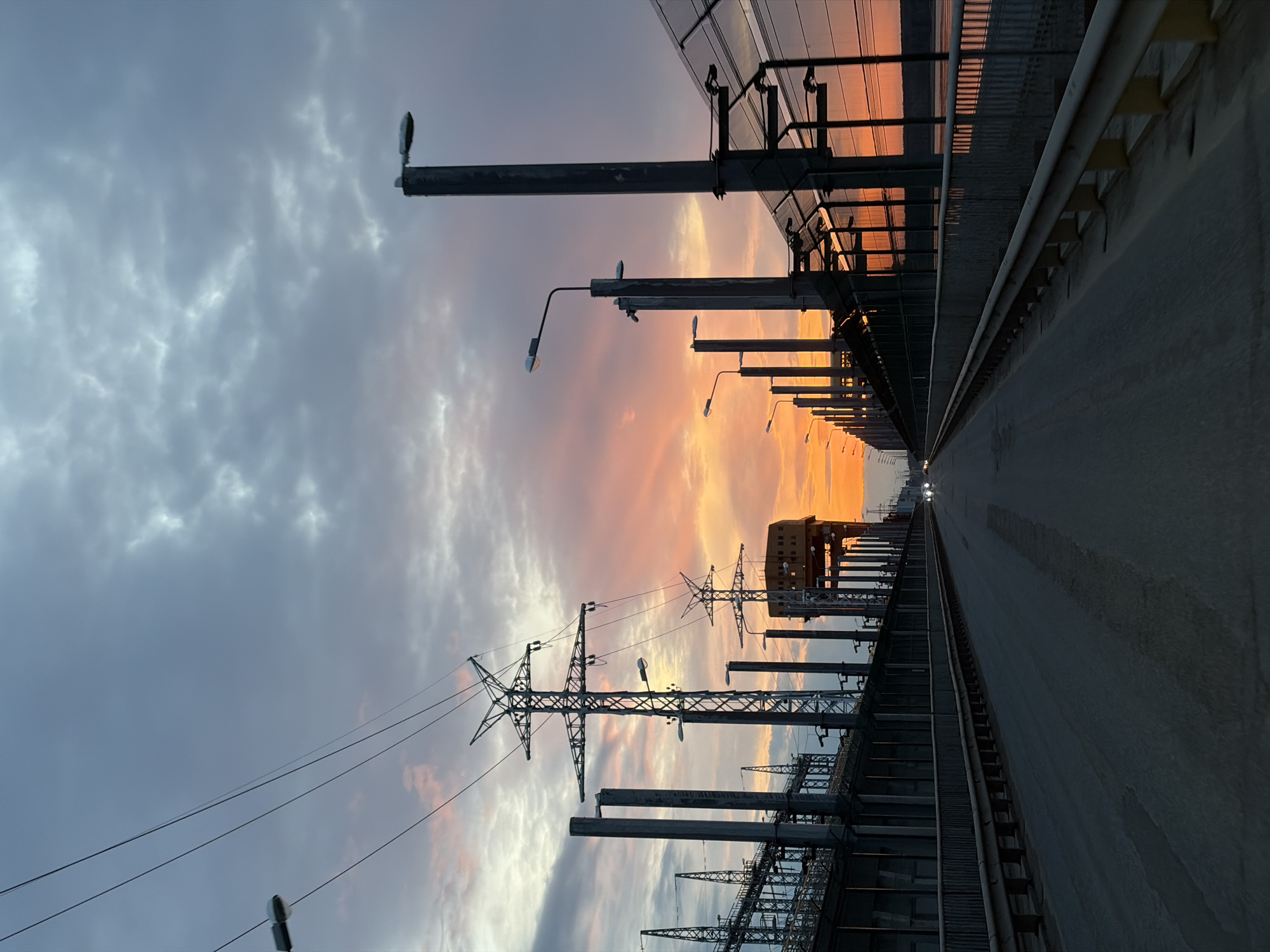 The image captures a view along a bridge in Chaikovsky, Russia, at sunset. The roadway stretches into the distance, flanked on both sides by a complex infrastructure of electrical power transmission equipment, including towers and substations. Streetlights line the bridge. The sky is a mix of clouds, with the setting sun casting orange and pink hues across the clouds. The roadway surface appears to be made of concrete. Two vehicles are faintly visible in the distance.