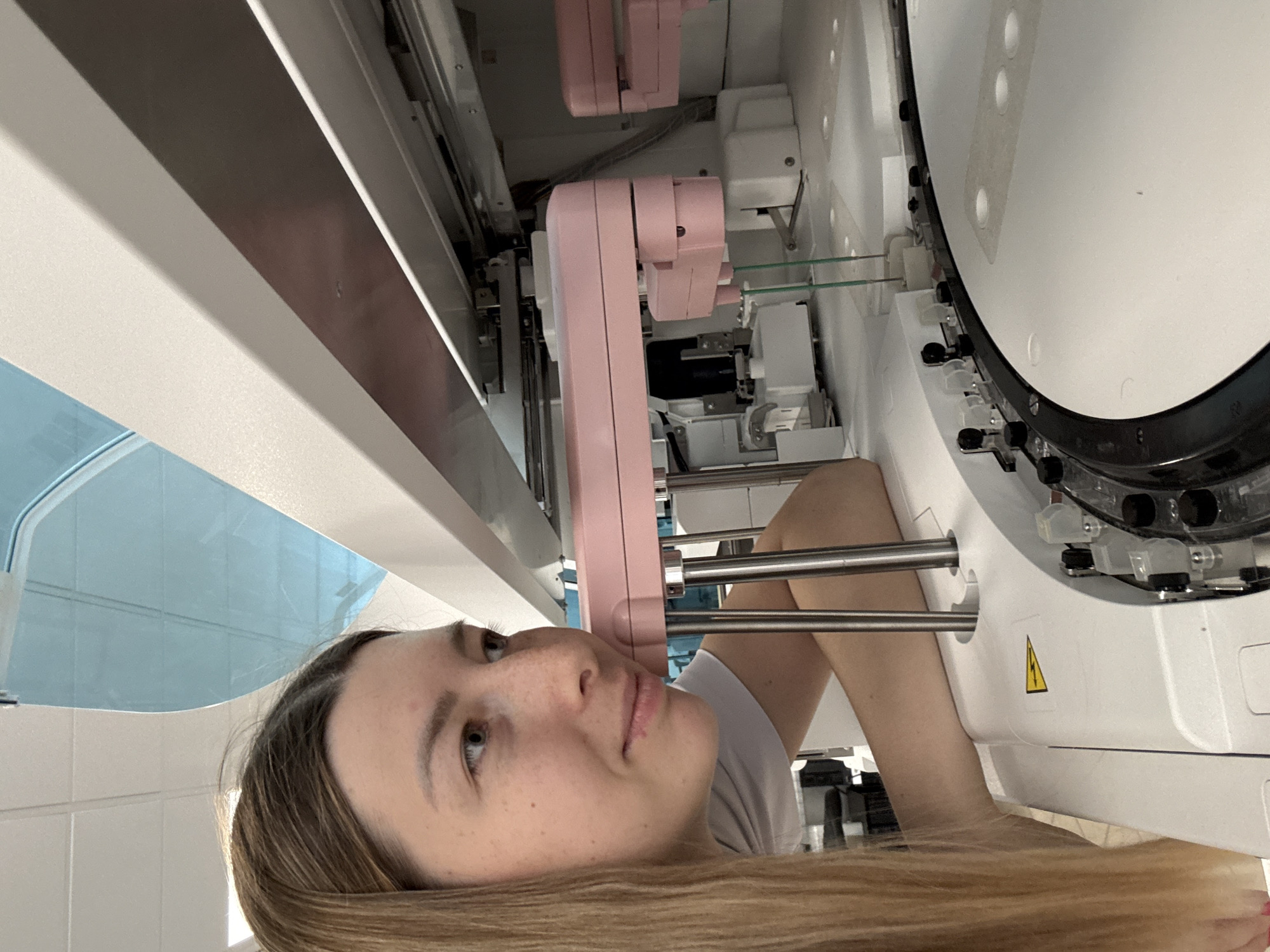 A young woman with long blonde hair and freckles leans towards a piece of laboratory equipment. She is wearing a white t-shirt. The equipment appears to be an automated analyzer with pink robotic arms. The scene takes place in a brightly lit room with white tiled ceilings. There is no information in the image to indicate the location is Moscow, Russia.