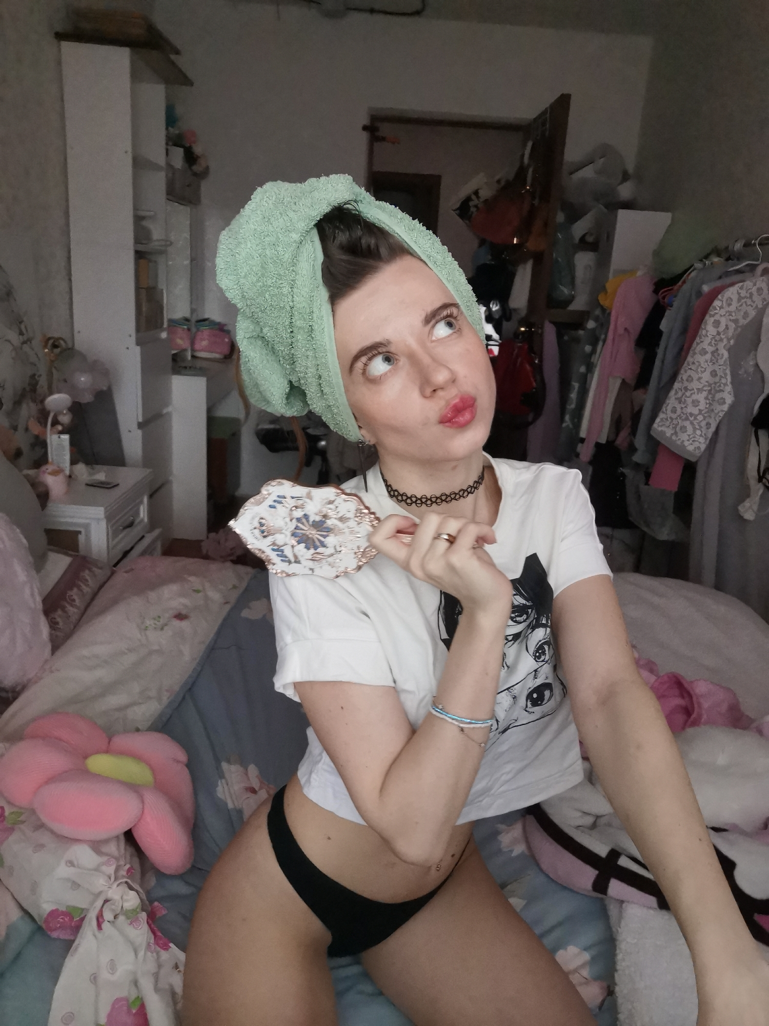 The image shows a young woman indoors. She is sitting on a bed with a light blue floral patterned bedspread, positioned in what appears to be a bedroom. The room's interior suggests it is in a state of disarray, with items such as clothing hanging on a rack in the background. The woman is wearing black underwear and a white t-shirt with an anime print. Her hair is wrapped in a light green towel, and she has a black choker around her neck. In her hand, she holds a decorative, oval-shaped object. She is gazing upwards with a pouting expression. A pink, flower-shaped pillow rests on the bed to her left. The location of the scene is not specified.