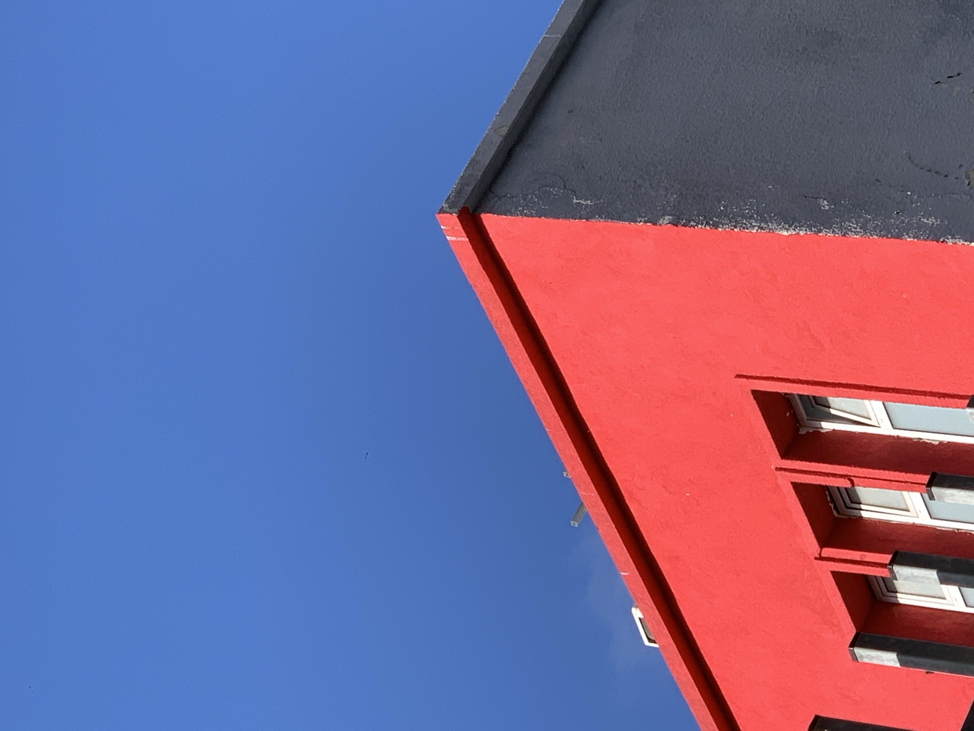 A low-angle shot captures the corner of a building under a clear blue sky. The building's corner is painted red, and the roof appears gray. Windows are visible along the red facade. The location is Tel-Aviv, Israel, but there are no people, subjects, actions, or interactions in the scene. The environment is clear and bright.