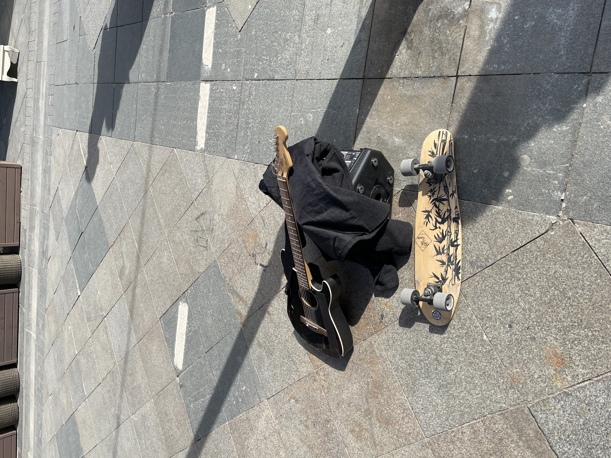 An outdoor scene on a paved area with stone blocks. There is a guitar leaning against a black bag, next to a small amplifier and a skateboard with a bamboo design. The location could be Moscow, Russia.