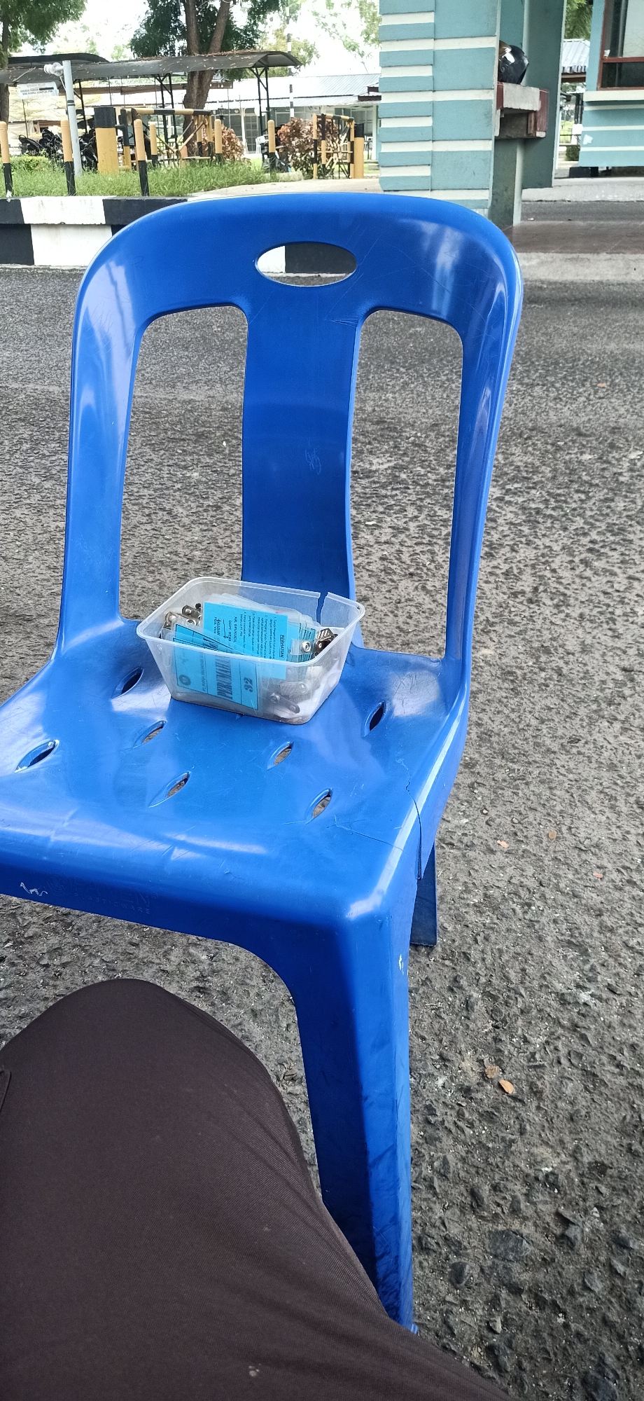 A blue plastic chair is positioned on an asphalt surface. On the seat of the chair rests a clear plastic container filled with blue identification cards, each featuring a metal clip. The chair and container are situated outdoors, with a building featuring a light blue and white striped facade visible in the background. Trees and other vegetation are also present. The bottom portion of the image shows the lap of a person wearing dark brown pants, suggesting they are seated. The scene appears to be in Tambontunong, Indonesia.