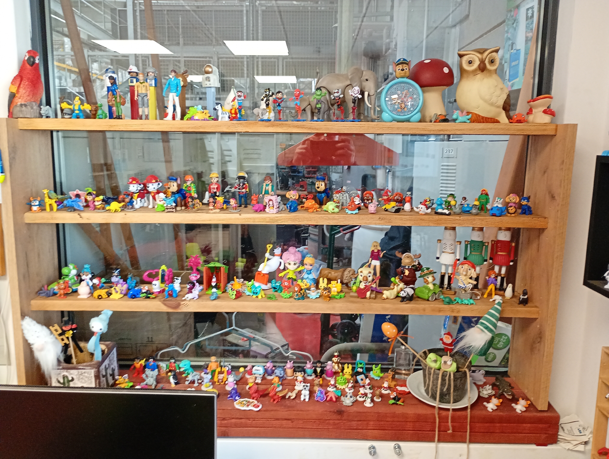A four-tiered wooden shelving unit, positioned behind a glass partition, displays a diverse and eclectic collection of hundreds of small figurines and toys. The shelves are packed with an assortment of characters, animals, and decorative objects, suggesting a personal hobby or a whimsical display.
On the top shelf, prominent items include a large red parrot figurine, various Playmobil figures (including an astronaut and explorers), a grey elephant, a red mushroom, and a beige owl. A blue Paw Patrol-themed clock sits beside the mushroom, topped with a character. Other small figures like Spiderman, cartoon characters, and small animals are interspersed.
The second shelf is largely populated by characters from the Paw Patrol franchise, alongside other animated figures, tiny cars, and miscellaneous plastic toys. The third shelf features a mix of fantasy characters, including Disney princesses, Stitch from Lilo & Stitch, several animals, and larger figures resembling nutcrackers.
The bottom surface of the unit holds an even more extensive collection of miniature figures. These include numerous small animals (dogs, cats, birds), various gnomes (one white fluffy, one blue ghost-like), cartoon characters, and several decorative items such as a small pot wrapped in string, an orange balloon on a stick, and a small white gnome with a green-striped hat.
Reflections in the glass partition show an indoor setting with industrial-style overhead lighting and metal framework, suggesting the location is likely an office, workshop, or similar commercial interior. Visible text includes "237" on a white box reflected on the second shelf, and "240" partially visible on a green item on the bottom shelf behind a gnome. The scene is set in Kabile, Bulgaria.