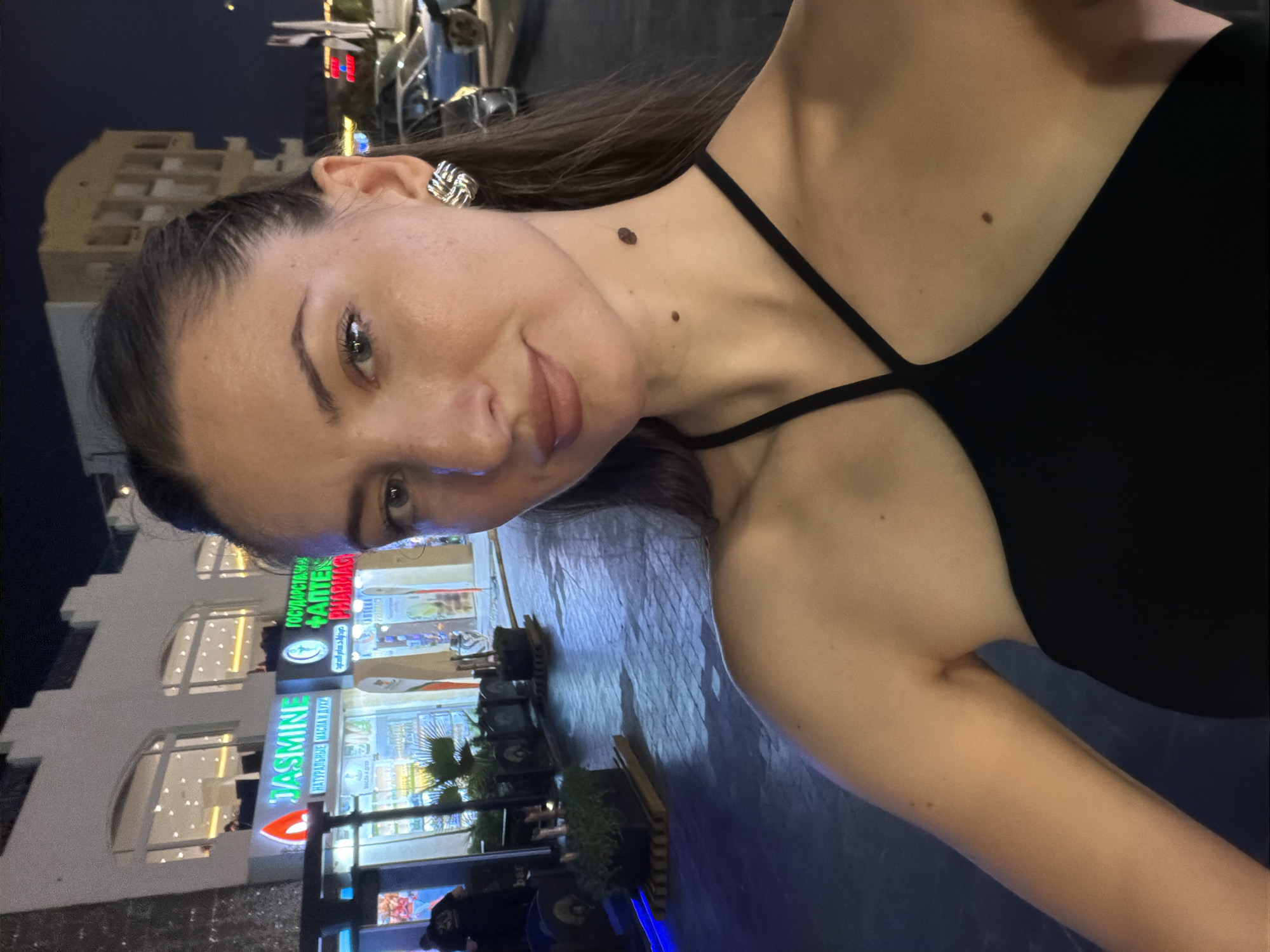 The image is a selfie taken at night on a street in Sharm El Sheikh, Egypt. The foreground features a woman with long, dark hair pulled back, wearing a black dress and large, silver earrings. She has two moles visible on her neck. In the background, there are storefronts with illuminated signs, including "JASMINE" and a pharmacy sign. There are also potted plants and parked cars on the street. The lighting suggests it is evening or night time.