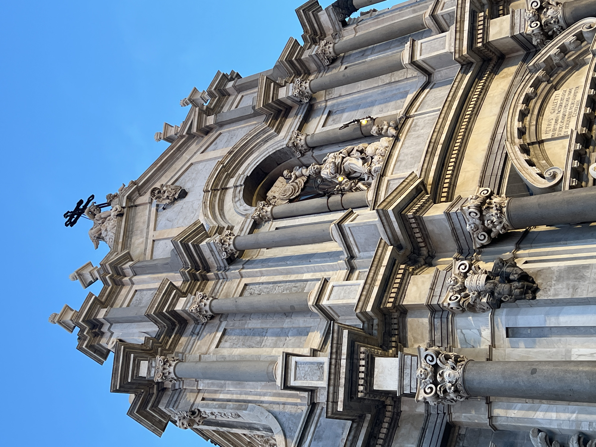 The image shows a low-angle view of the facade of the Catania Cathedral, also known as the Duomo di Catania, located in Piazza del Duomo, Catania, Italy. The baroque architecture features intricate stonework, columns, and sculptures. A prominent cross is visible at the top of the facade against a clear blue sky. In the center of the facade, within an alcove, is a sculptural group depicting figures, including what appears to be a seated woman holding a child, surrounded by cherubs and other smaller figures. Below this sculpture, an inscription is visible on a stone plaque. The stonework is a light gray marble with darker gray columns and accents. The sunlight casts shadows, highlighting the architectural details. No people are visible in the scene.