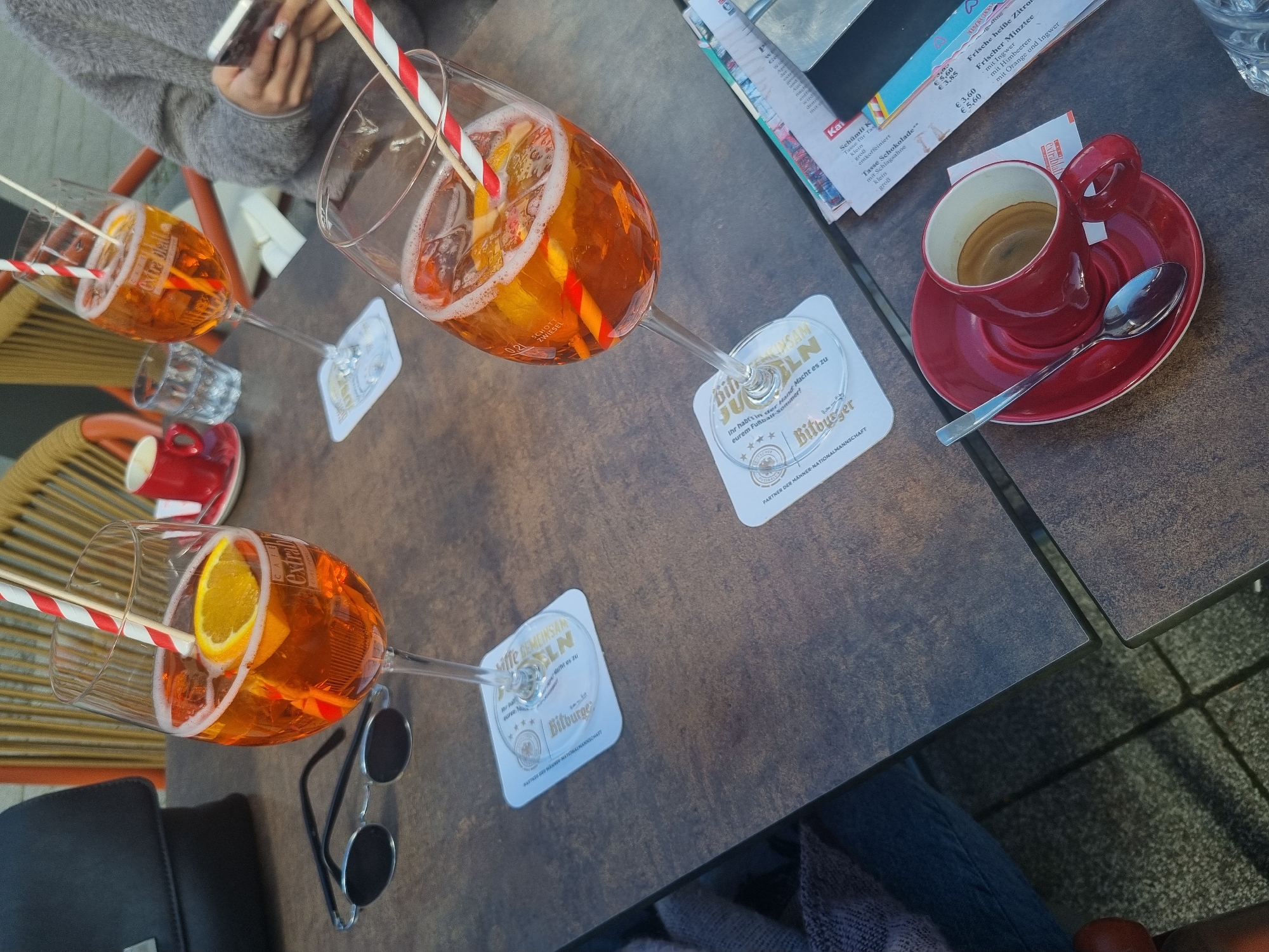 The scene captures an overhead view of a table in what appears to be a restaurant or café in Saarbrücken, Germany. The table is constructed of dark brown planks. At least three glasses of Aperol Spritz with straws are on the table. They are resting on branded coasters with "Bitburger" written on them. There is also a red espresso cup and saucer with a spoon on the table, along with a printed menu or price list. In the bottom left of the frame, a pair of sunglasses is visible near one of the Aperol Spritz glasses. Parts of people are visible; someone is holding a phone in the upper left corner, and the clothes of patrons sit at the table's edge. The setting suggests an outdoor dining area on what seems to be a partly cloudy day.