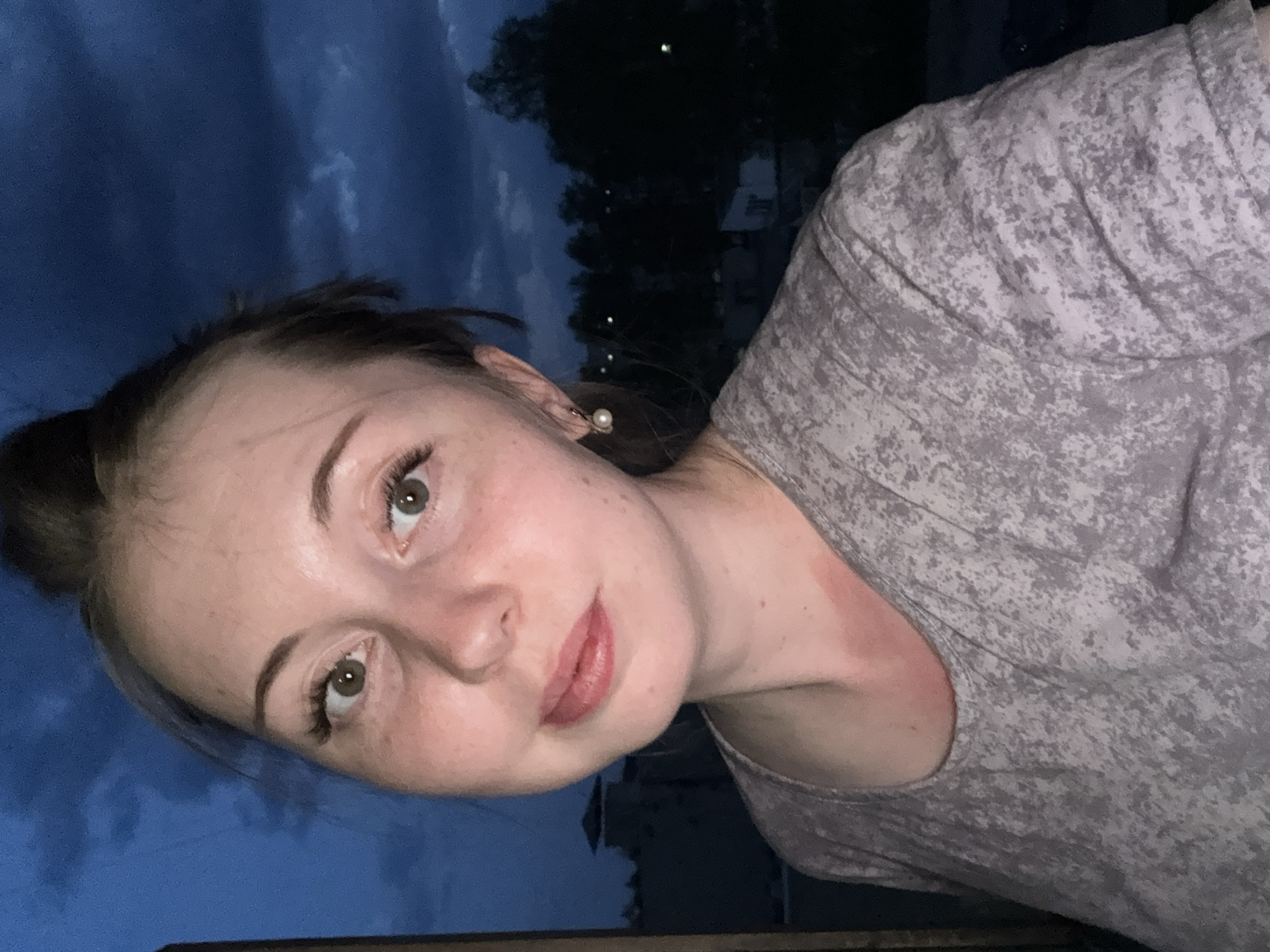The image shows a woman in a grey mottled t-shirt, posing for a selfie outdoors. She has brown hair pulled back, fair skin, and is wearing pearl earrings. The background suggests it is evening, with a dark sky, trees, and buildings visible. The location is Unknown, Unknown.