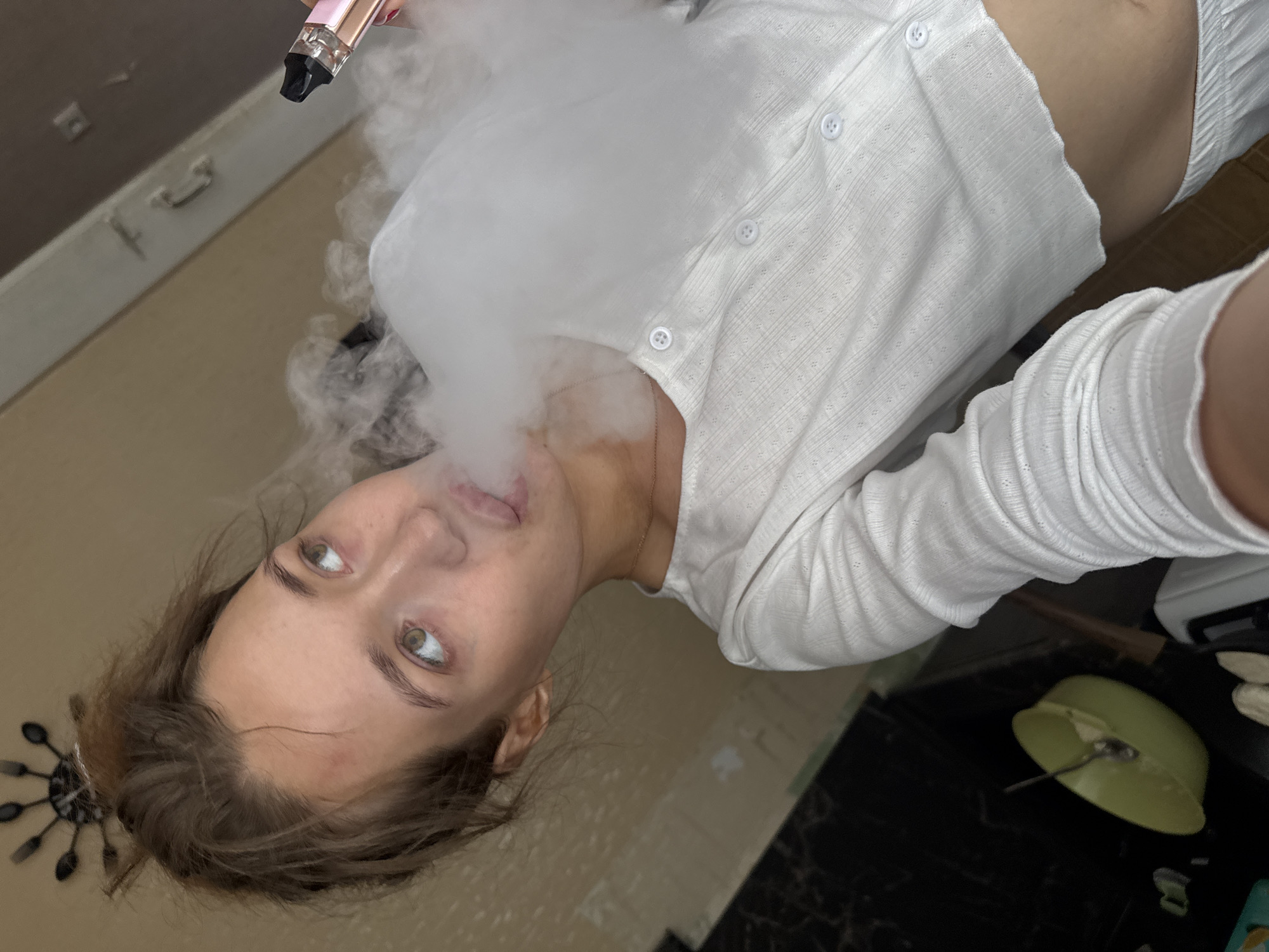 A young woman is shown in a selfie, exhaling a cloud of vape smoke. She is wearing a white, long-sleeved, button-up top. Her hair is pulled up loosely. Her eyes are looking upwards and to the right. The background appears to be an indoor setting with a textured wall and some kitchen elements visible, such as a bowl with a spoon in it.