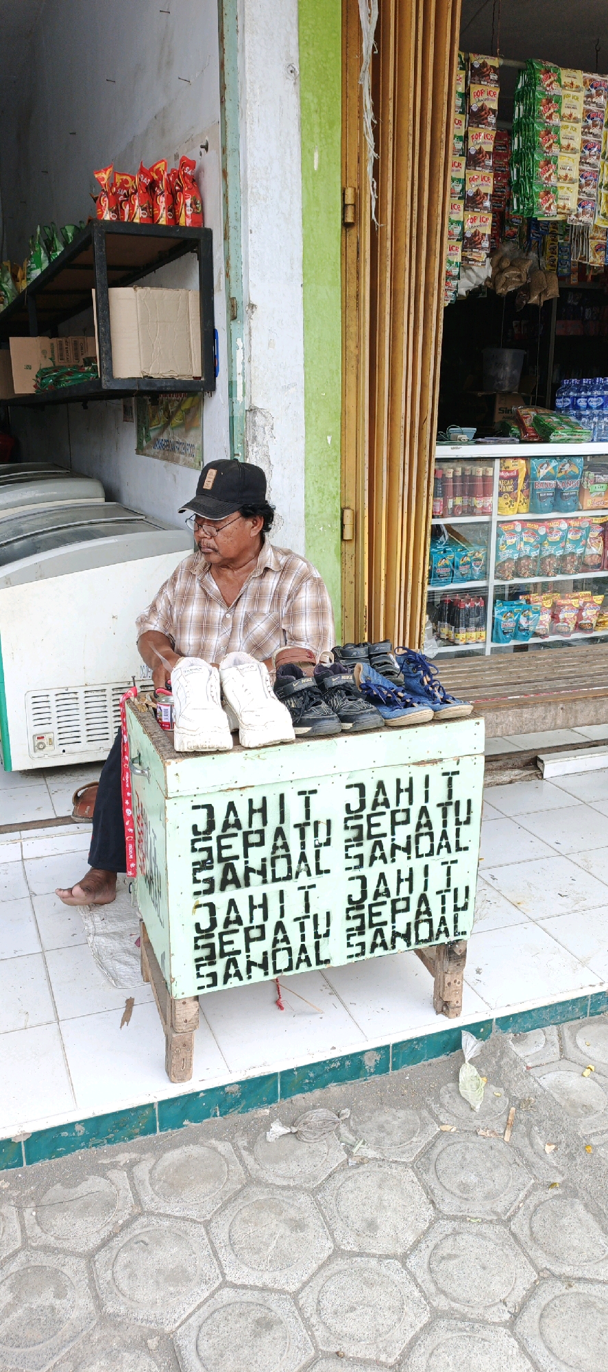 A man wearing a plaid shirt, glasses, and a black baseball cap sits behind a light green wooden cart, which has black lettering that reads "JAHIT SEPATU SANDAL" (meaning "Shoe and Sandal Stitching") repeated on its front. A pair of white sneakers and several pairs of black and blue sandals are displayed on top of the cart. The man is looking down at the cart. Behind him, a shop entrance is visible with a brown accordion-style door and shelves stocked with various packaged goods. The shop is situated on a tiled sidewalk in front of a building with a light green and white facade. The ground in front of the shop is paved with hexagonal concrete tiles, and there is some debris on the ground. The overall scene suggests a small street-side business in Kasiyan, Indonesia, likely a cobbler's stall.