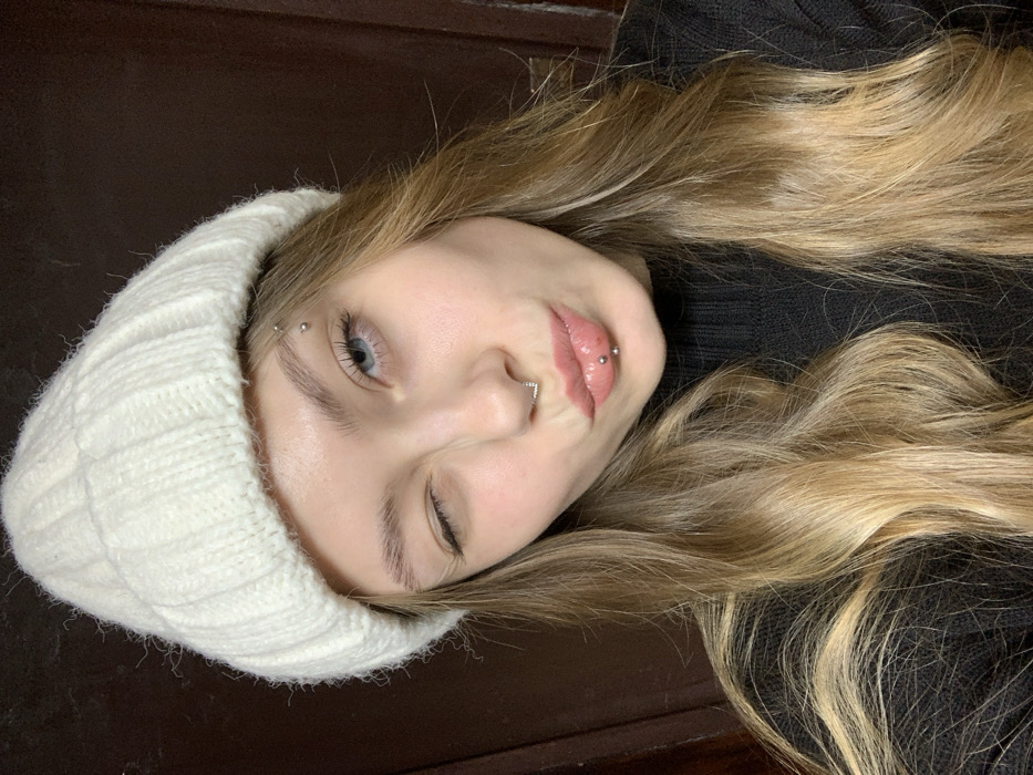 A young woman with blonde hair and blue eyes is wearing a white knit hat. She is winking and has multiple piercings, including eyebrow, nose, and lip piercings. She is wearing a black sweater, and the background appears to be a dark brown wall. The photo is taken at an angle, with the woman tilting her head.