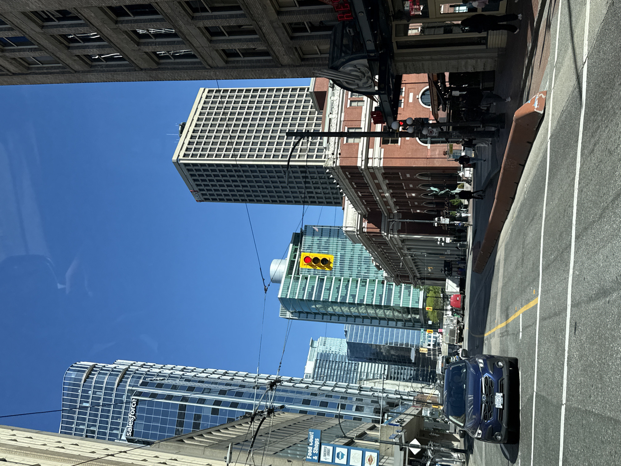 The image shows a street scene in downtown Vancouver, Canada. The perspective is from inside a vehicle looking out onto a street with several tall buildings on either side. On the right is a brick building with rounded windows on the top floor, accompanied by a number of people walking on the sidewalk. The adjacent road is marked with white lines, indicating lanes, and a yellow line dividing the street. On the right lane, a dark blue SUV is parked. A traffic light with a yellow light visible is suspended above the street. Across the street, a building displays the "salesforce" sign. The sky is clear and blue.