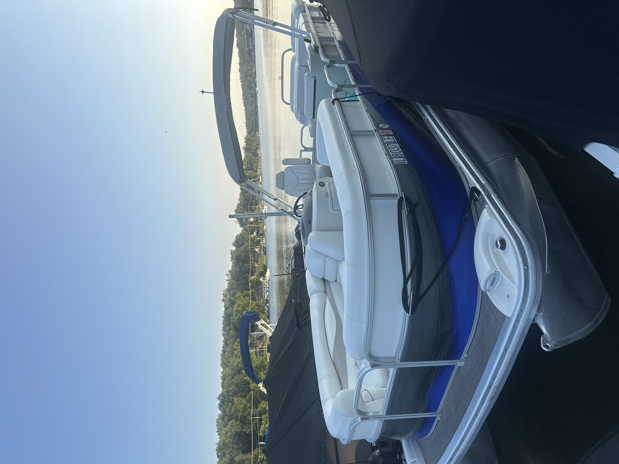 A pontoon boat, identified by the registration "IL 8037 LE", is docked on a body of water, likely a lake or river, in Algonquin, Illinois, USA. The boat features white upholstery and a blue and gray exterior. It is positioned alongside other boats, some of which are covered with black tarps. The background shows a tree-lined shore with houses and a clear blue sky. The scene appears to be set during daytime, with the sun casting a gentle light.