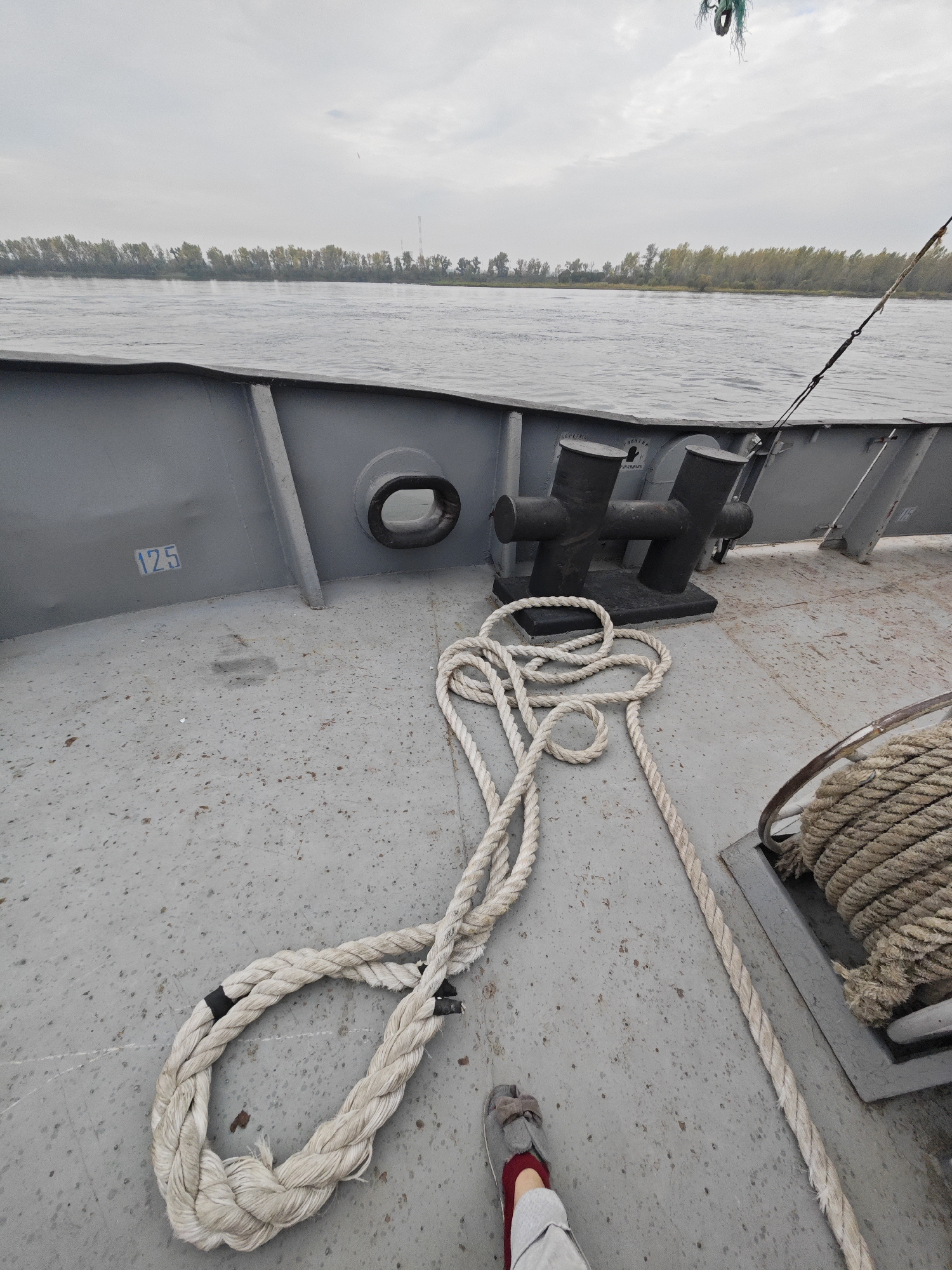 The image shows a deck of a gray boat with a thick white rope coiled on the surface. In the background, a calm body of water is visible, with a tree-lined shore in the distance under a cloudy sky. A metal bollard, a common fixture on boats and docks for securing ropes, is also present on the deck. A person's legs are visible in the lower right of the frame, wearing gray pants and red socks with gray slippers. The scene appears to be a still moment on the water, possibly in the vicinity of Ермолаево, Russia.