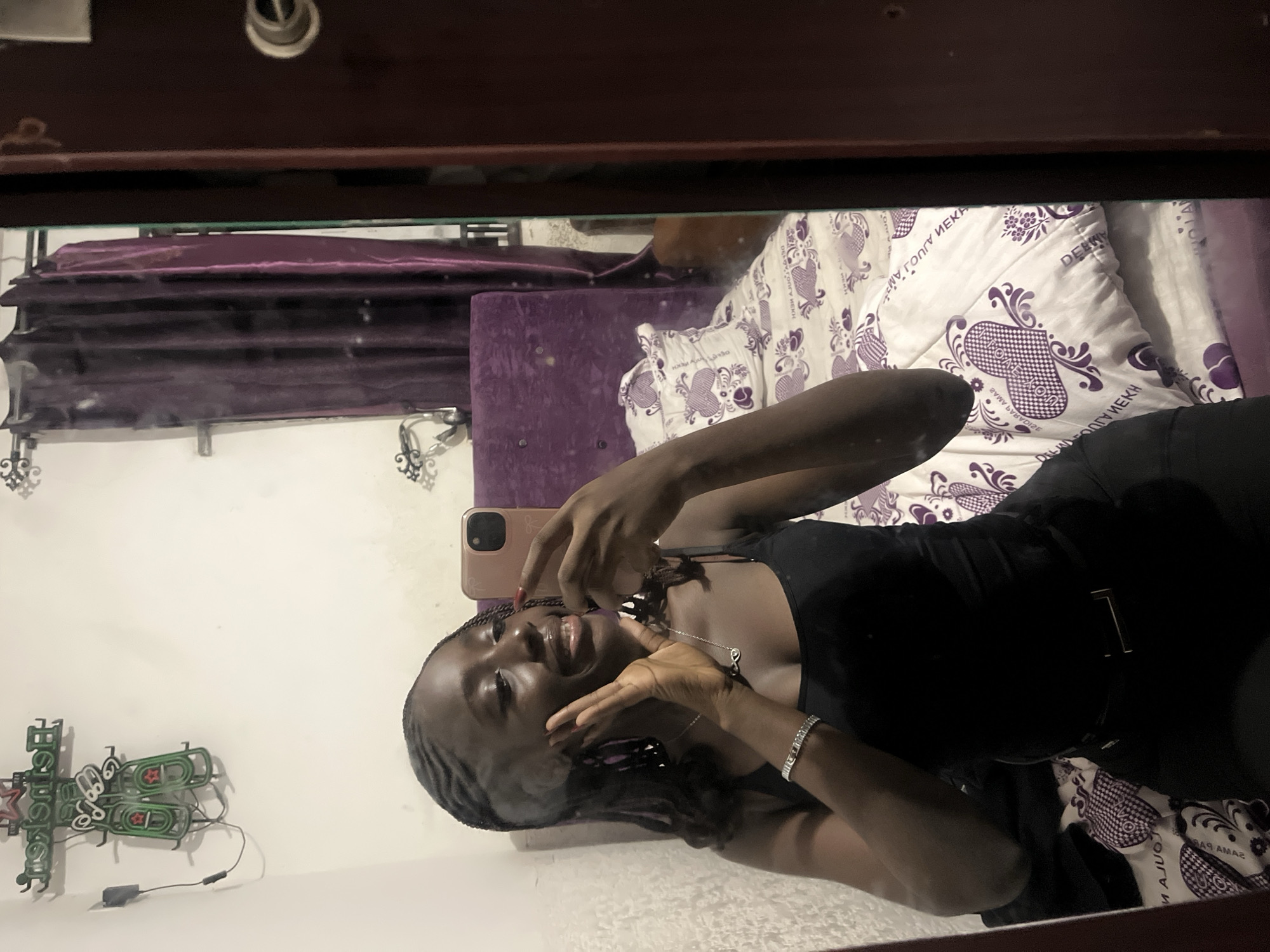 A young woman is reflected in a mirror, holding a smartphone as if taking a selfie. She has dark skin, black braided hair, and is wearing a black top with a belt and a silver bracelet and necklace. Her hands are near her face. Behind her, reflected in the mirror, is a bed with purple and white patterned bedding and a velvet headboard. On the wall to the left, a neon sign in the shape of beer bottles is visible. The background suggests an indoor bedroom setting. There is no information in the image to suggest the location is Abuja, Nigeria.