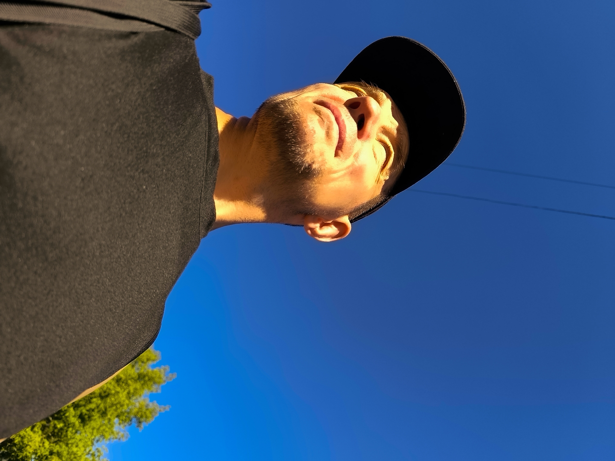 A low-angle, close-up shot features a young person wearing a black t-shirt and a black baseball cap. The individual's face is illuminated by sunlight, casting shadows that highlight facial features such as the nose, lips, and stubble. The person appears to be looking upwards towards a clear, bright blue sky. In the lower left corner of the frame, the upper branches of a vibrant green tree are visible, suggesting an outdoor setting. The background is dominated by the expansive sky, with faint, dark horizontal lines that may indicate power lines. The image has a dynamic, upward perspective.
