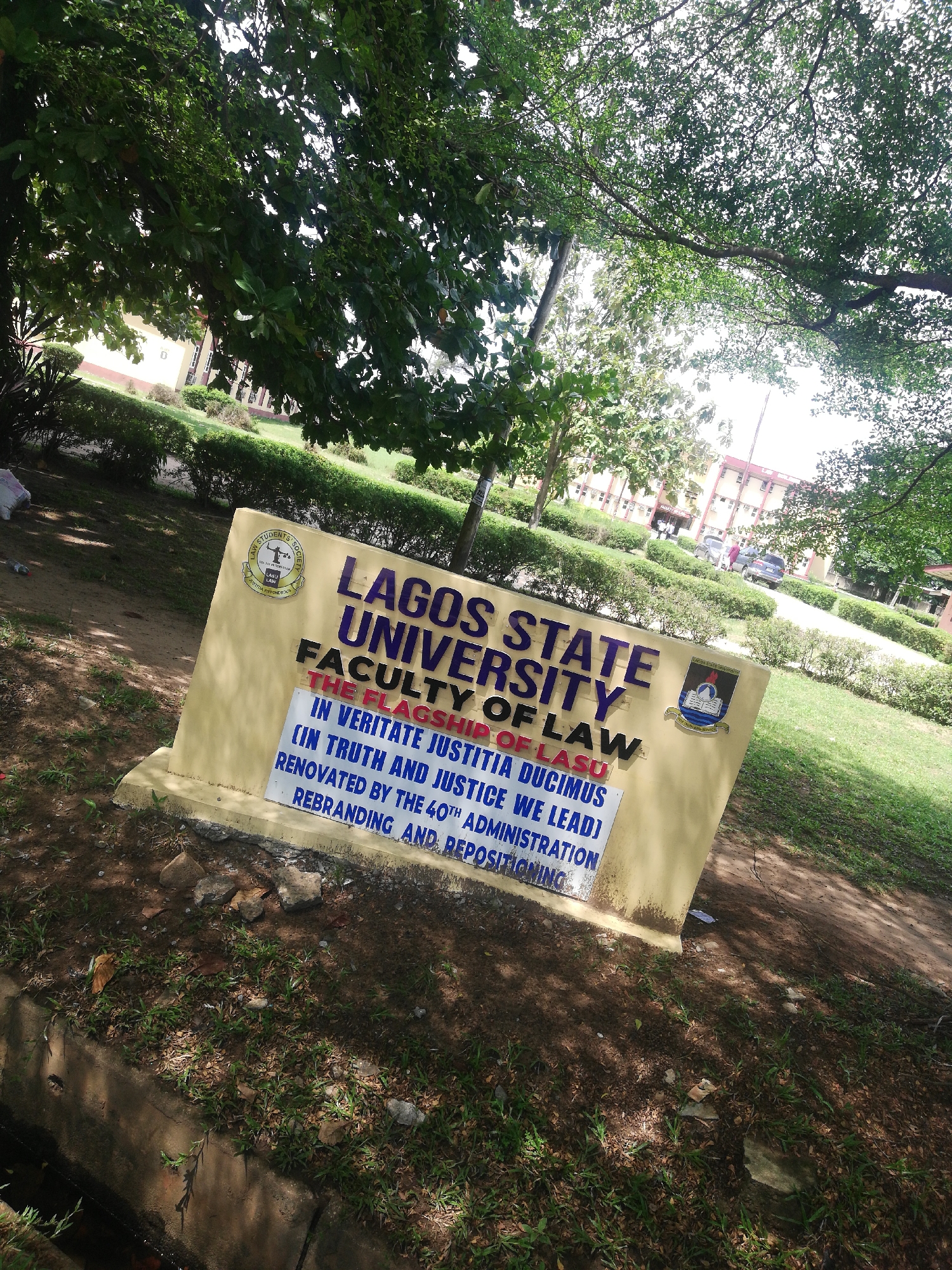 This outdoor, daytime photograph captures a sign marking the Faculty of Law at Lagos State University (LASU) in Lagos, Nigeria. The sign, painted in a beige or light yellow color with blue lettering, is prominently displayed in the lower-middle of the frame, angled slightly to the right. Above the main text on the sign, to the left, is a circular emblem, partially obscured but showing "LAGOS STATE UNIVERSITY" and "STUDENTS' SOC." (likely "Students' Society"). To the right of the main text, another crest is visible.
The text on the sign reads: "LAGOS STATE UNIVERSITY," "FACULTY OF LAW," "THE FLAGSHIP OF LASU," "IN VERITATE JUSTITIA DUCIMUS," "(IN TRUTH AND JUSTICE WE LEAD)," "RENOVATED BY THE 40TH ADMINISTRATION," and "REBRANDING AND REPOSITIONING."
The setting is a university campus, characterized by lush green foliage. A large tree canopy dominates the upper portion of the image, providing dappled sunlight and shadows. Behind the sign, there is a neatly trimmed lawn with manicured hedges. In the background, university buildings with reddish-brown roofs and light-colored walls are visible, along with what appear to be parked cars and distant figures, suggesting general campus activity. Below the sign, a concrete drainage ditch runs along the bottom edge, with scattered leaves, small rocks, and patches of grass on the ground. The bright, natural lighting indicates a clear, sunny day.