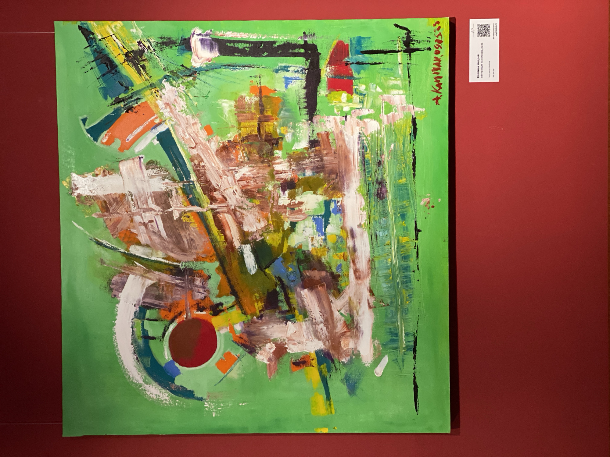 A green abstract painting hangs on a red wall. The painting features a variety of brushstrokes and colors including green, brown, white, orange, and black. The painting also features a red circle near the bottom left corner. A small white label with black text and a QR code is affixed to the wall to the right of the painting. The painting is signed in the upper right corner. The artwork is on display at a gallery in Moscow, Russia.