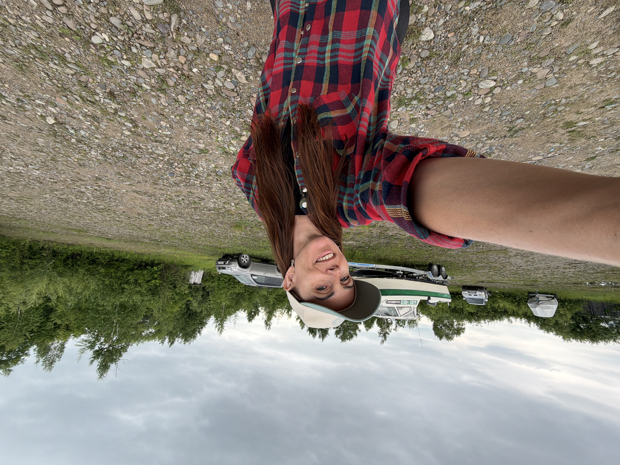 In Talakan, Russia, a woman with long, brown hair, wearing a red and blue plaid shirt and a tan baseball cap, takes a selfie outdoors. She is smiling at the camera. Behind her, a white and green boat on a trailer is parked on a gravel lot. In the background, several vehicles and a densely wooded area are visible under a cloudy sky. The ground is a mix of gravel and patchy grass.