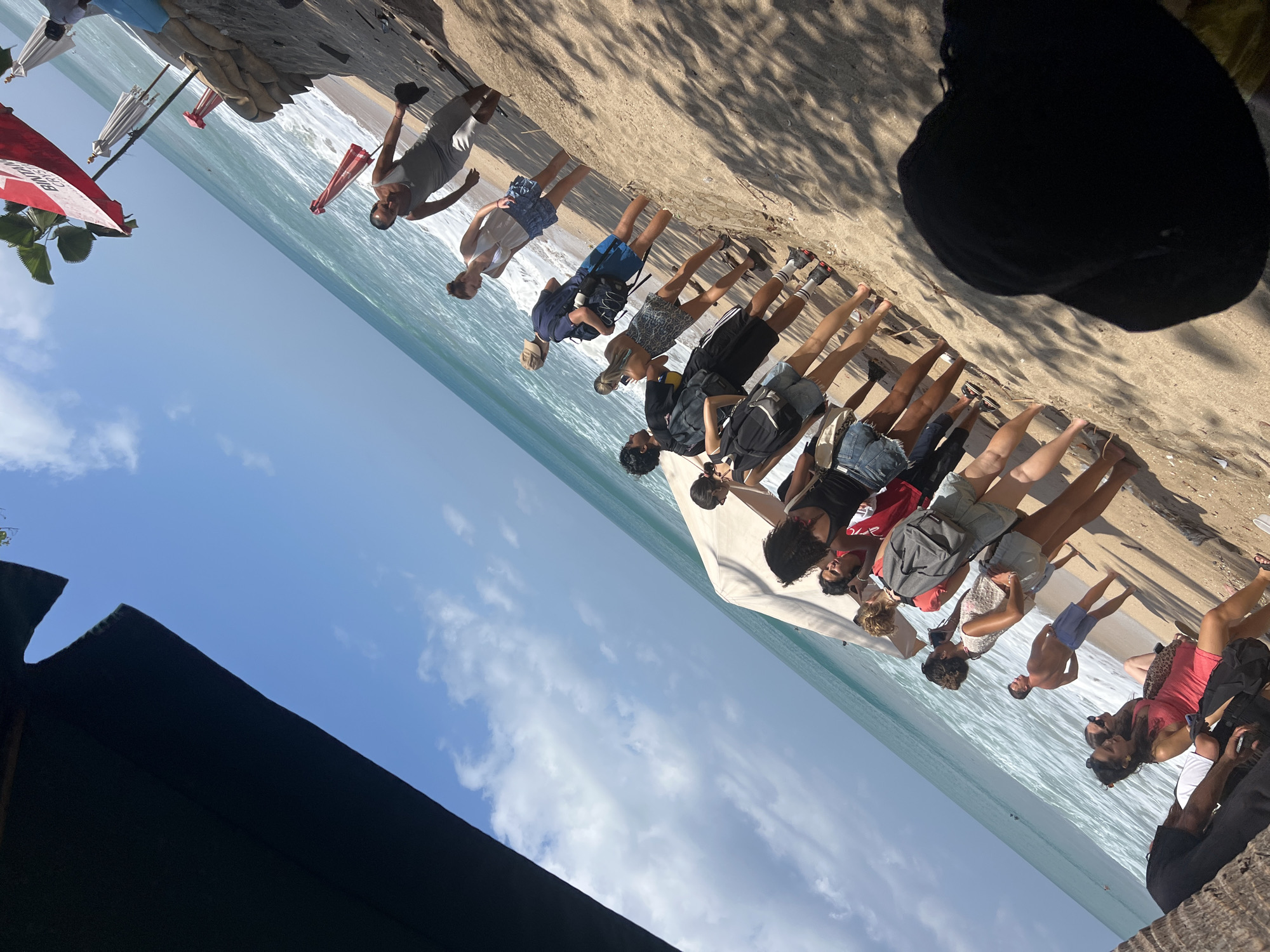 A group of people are gathered on a sandy beach in Kuta, Indonesia, with the ocean and sky in the background. The image is taken from a low angle, partially obscured by a dark, possibly thatched, overhead structure. The people are standing and sitting, some are looking at the ocean, others are interacting with each other.
In the foreground, a person wearing a black hat is seated, facing away from the camera. To their left, a couple is seated on the sand. The woman is wearing a pink dress, and the man is wearing dark shorts.
Moving further back, a line of people are standing on the beach, facing towards the ocean. They are dressed in casual beach attire, including shorts, t-shirts, and tank tops. Some have backpacks. The sand is light brown, and the ocean has light blue waves.
In the background, there are beach umbrellas, some of which are red and white, and others are white. There are also sandbags stacked along the edge of the beach. The sky is blue with scattered white clouds. The sun is shining brightly, casting shadows on the sand. The overall atmosphere appears to be relaxed and social.