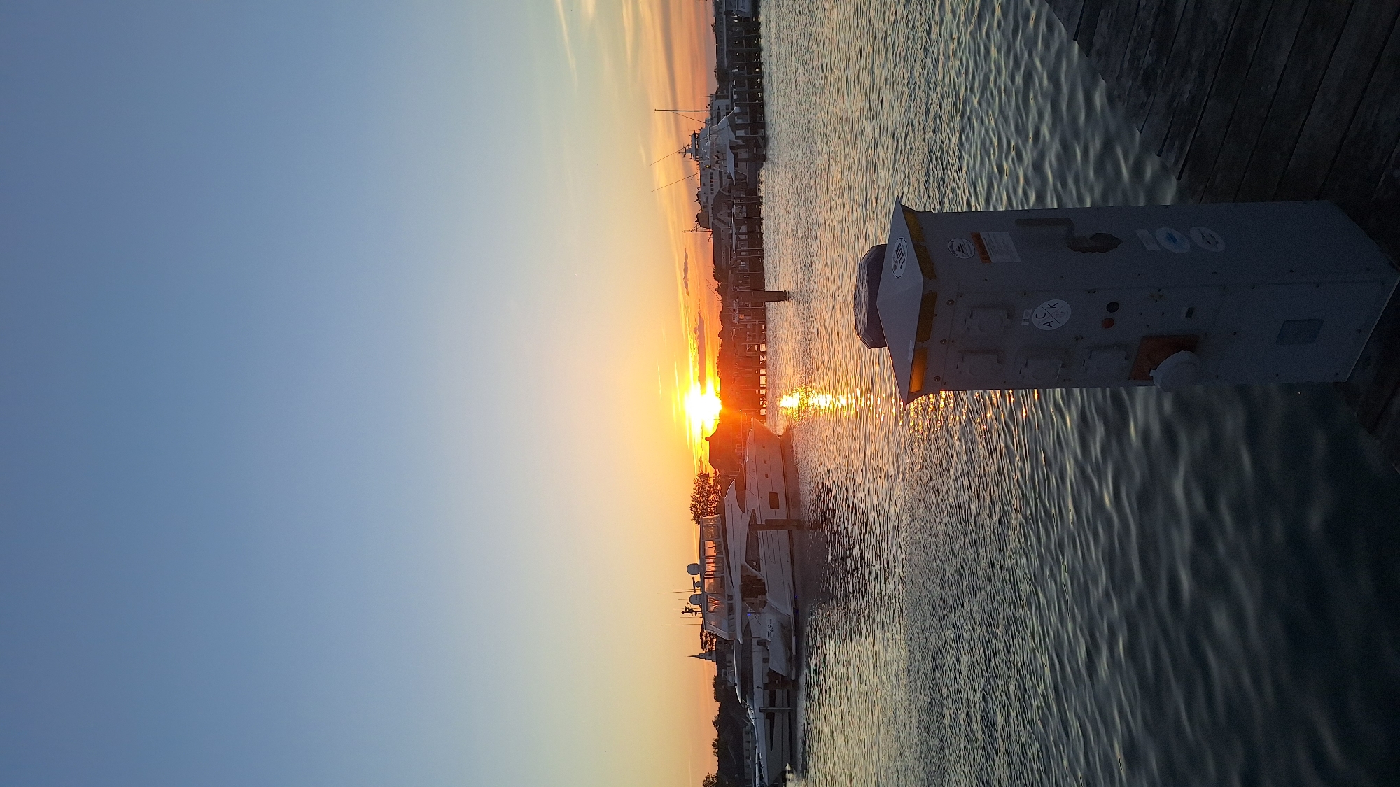 An image captures a serene sunset over a harbor in Nantucket, Massachusetts, USA. The sky transitions from a clear, soft blue at the top to warmer hues of orange and yellow near the horizon. The setting sun, a bright orb, casts a shimmering reflection on the water, creating a path of golden light that stretches towards the viewer.
Several white yachts of varying sizes are docked, their sleek forms silhouetted against the colorful sky. Some appear to be luxury vessels, with multiple decks and railings. In the background, a wooden pier extends into the water, lined with more boats and buildings, including what appears to be a church steeple. The water itself is calm, with gentle ripples reflecting the light and the surrounding scene.
In the foreground, on the right side of the image, a grey electrical pedestal stands on a wooden dock. This utility box has multiple outlets and various stickers, including one circular sticker with the letters "ACK" and a smaller, round sticker with a depiction of a boat and the number "100". The wooden planks of the dock are visible beneath the pedestal, showing signs of weathering. The overall atmosphere is one of tranquility and the peaceful end of a day.
