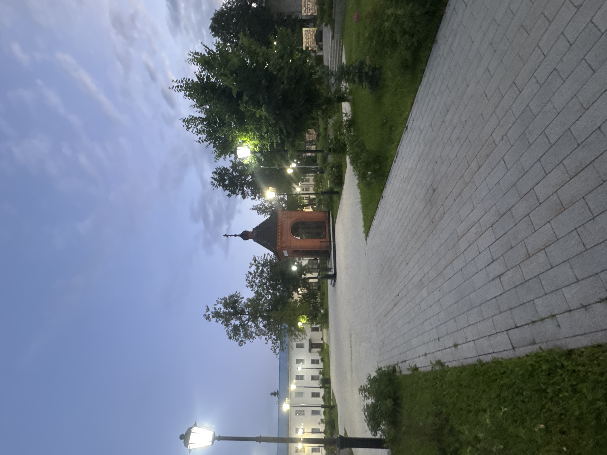 This is a nighttime exterior shot of a paved courtyard area in the city of Vereya, Russia. In the foreground, a wide, gray brick pathway leads into the scene, bordered by a strip of green grass. Several ornate street lamps with glowing bulbs line the pathway.
To the left of the pathway, a long, white building with numerous windows and a blue roof is visible. In the center of the courtyard, a small, red brick chapel or shrine with a dark, domed roof and a cross on top stands prominently.
Behind the chapel, a variety of green trees create a natural backdrop. The sky above is a deep twilight blue with wispy clouds. The scene appears serene and well-maintained, suggesting a public or religious space. There are no people or animals visible in the image.