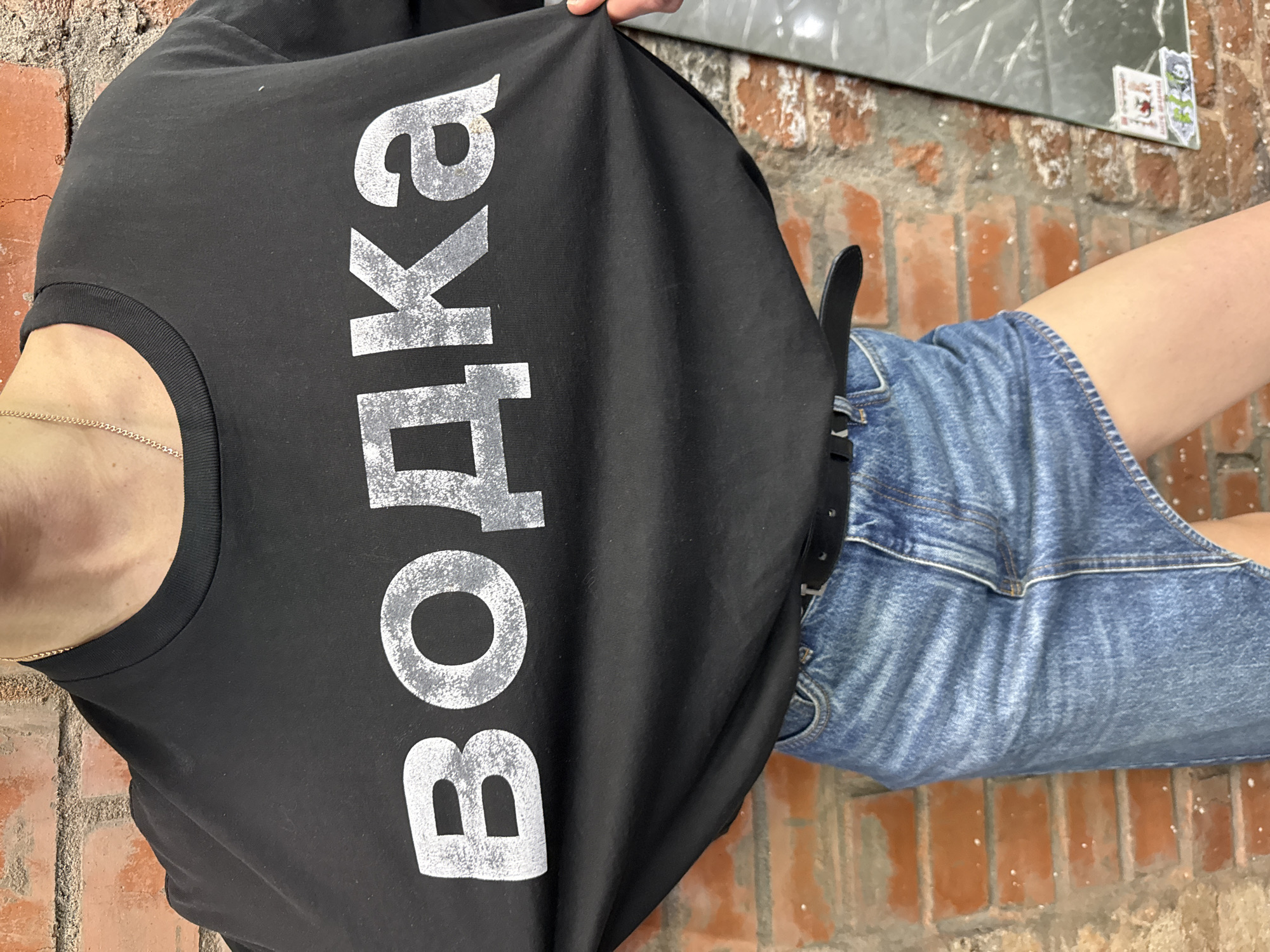 A woman is wearing a black t-shirt with the word "ВОДКА" printed vertically in large, distressed white lettering. She is also wearing a blue denim skirt with a black belt. The background consists of a brick wall, part of which is whitewashed, and a mirror with stickers on it. The location is likely Moscow, Russia due to the Cyrillic text on the shirt.