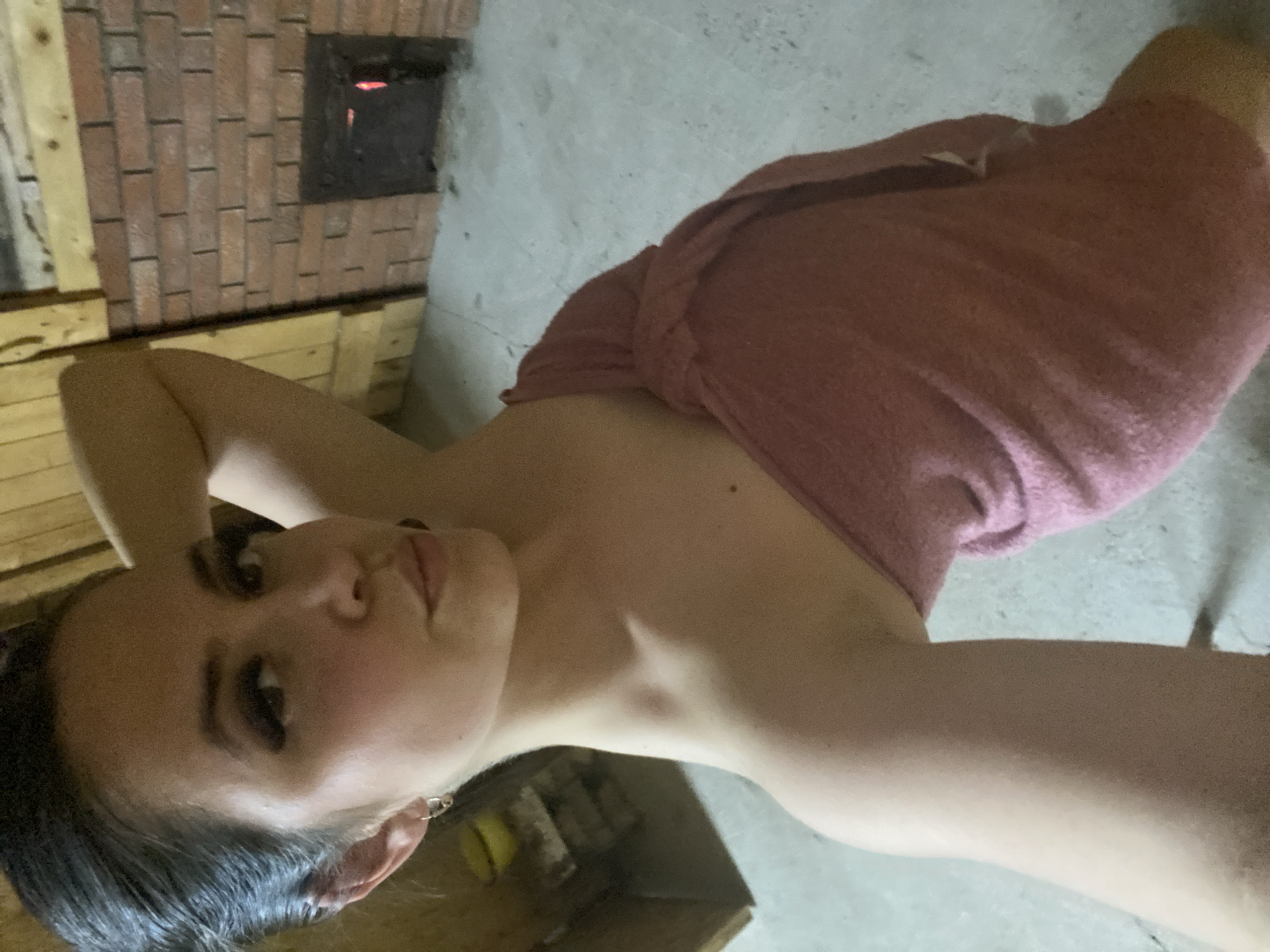 A woman wrapped in a pink towel is positioned in what appears to be a sauna or bathhouse. Behind her is a brick wall with a small firebox opening. Wooden planks line the wall below the bricks. The floor is concrete. The location, Zhigalki, Russia, is outside the scope of what can be determined from the visual information provided.