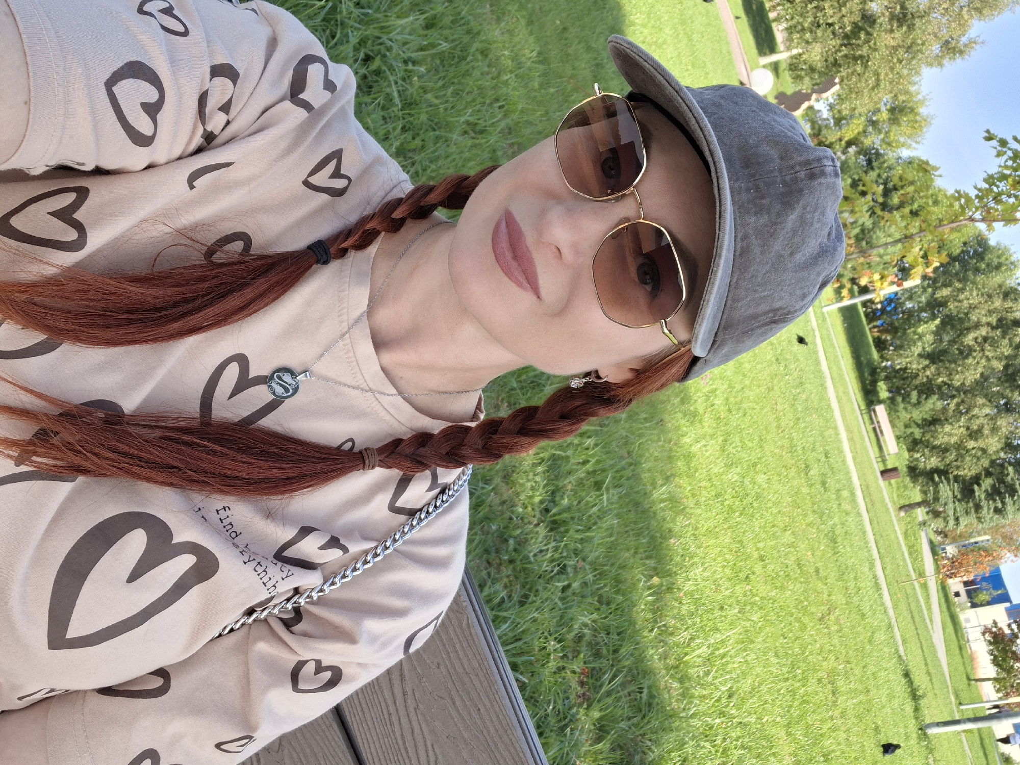 A woman wearing a gray baseball cap and gold-rimmed sunglasses sits on a park bench outdoors, her head turned to the right to face the camera. She has auburn hair styled in two braids and is wearing a light-colored shirt with a pattern of dark hearts. A silver chain necklace with a circular pendant is visible around her neck. The background shows a grassy park with trees and a blue sky. The location is identified as Myadzyel, Belarus, and the scene appears to be a casual outdoor setting on a sunny day.