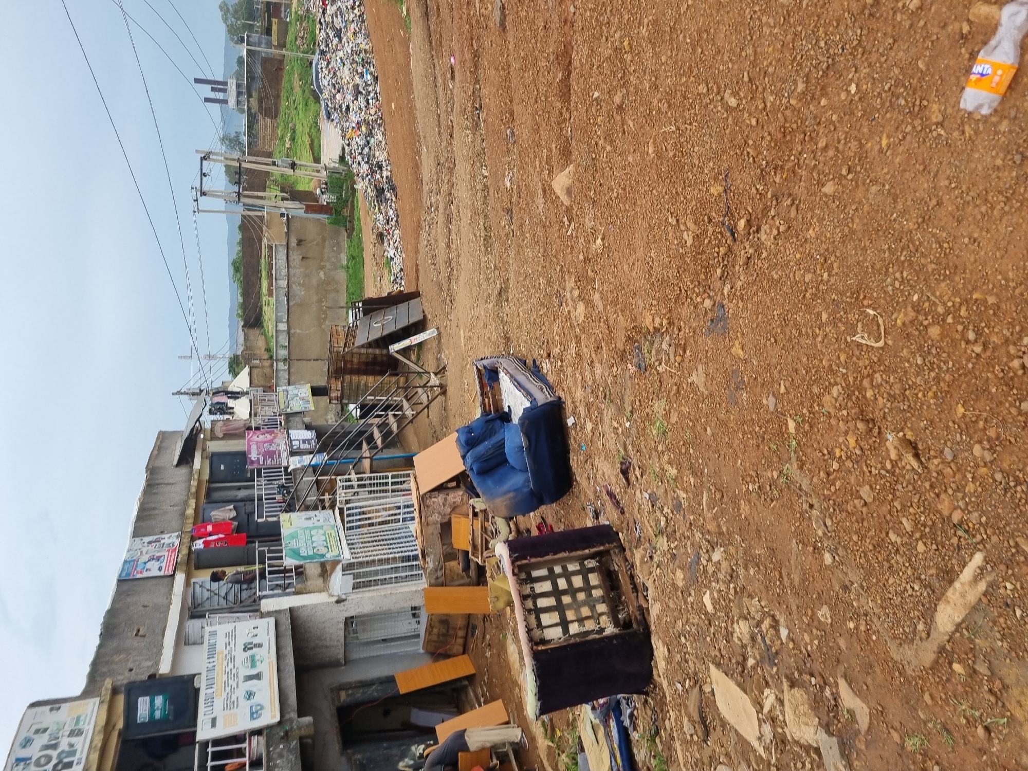 The image depicts an outdoor scene in Jos, Nigeria, characterized by urban decay and neglect. In the foreground, discarded furniture, including a dark blue sofa and the frame of an armchair, lies on a dirt road covered in small stones and gravel. To the left, a multi-story building houses several small shops with open entrances. Signs for "CLASSIC PRINTING & PRODUCTION" and other services are visible. A few individuals are seen near the building, one of whom appears to be an employee or customer.
The middle ground reveals more scattered debris and what looks like remnants of construction or renovation materials, such as wooden boards. Further into the background, a large accumulation of trash and refuse is piled high, indicating a significant waste disposal issue. Beyond the landfill, a residential or commercial area with low-rise buildings and green vegetation can be observed, under a cloudy sky. Electrical poles and overhead wires are present, suggesting ongoing utility infrastructure. The overall impression is one of an unkempt urban environment with visible signs of poverty and a lack of adequate waste management.