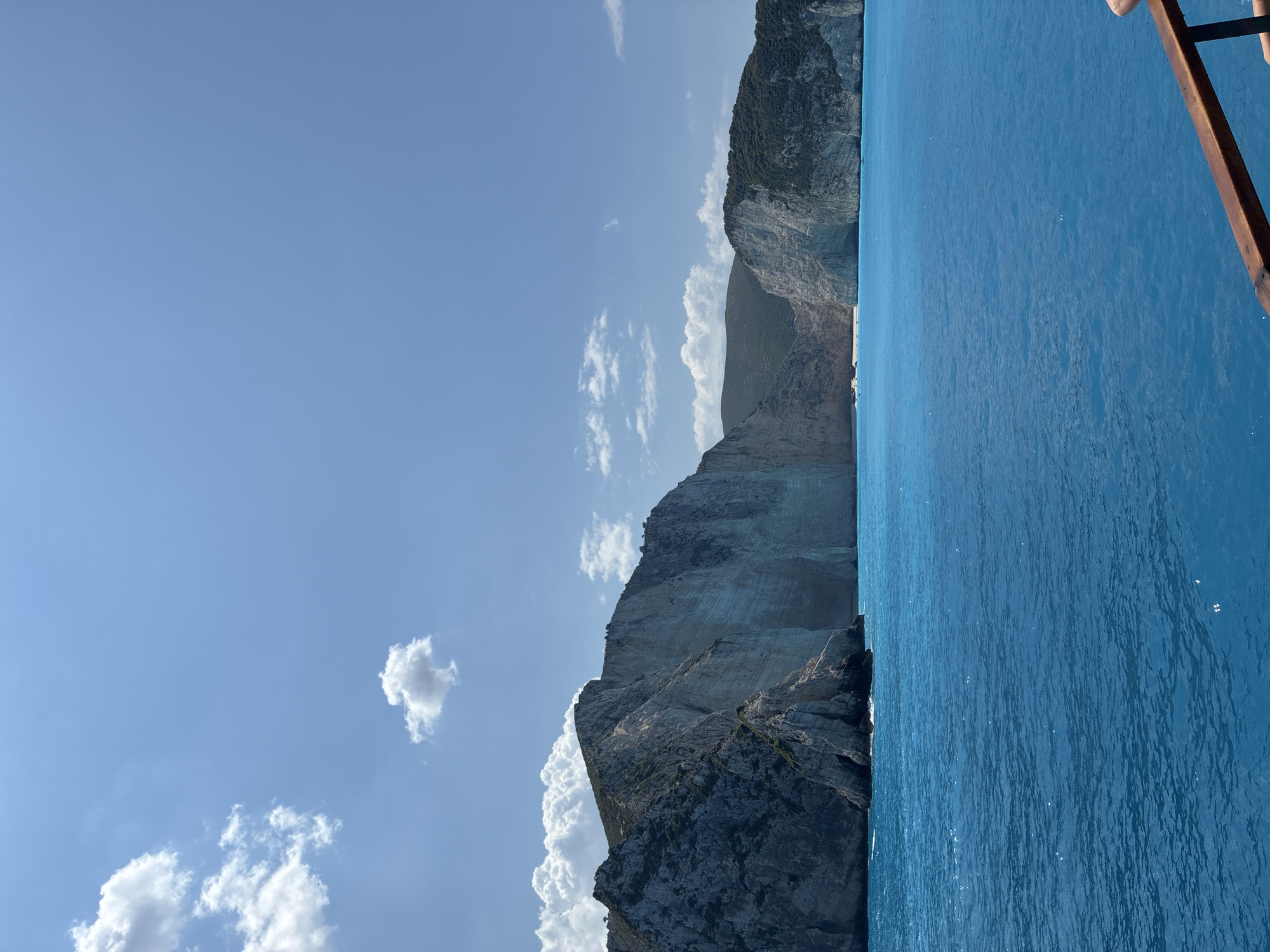 The image captures a scenic view of the coast of the Elaties Municipal Unit, Greece. Towering limestone cliffs dominate the background, with patches of green vegetation clinging to their slopes. The cliffs are rugged and striated, showcasing the geological formations of the region. In the foreground, the bright turquoise waters of the sea extend outwards, with small white waves and ripples visible on the surface. The sky is a clear blue, dotted with a few fluffy white clouds, suggesting a sunny day. The lower right corner of the image shows a wooden railing, implying the photograph was taken from a boat or a similar elevated vantage point. No people are visible in the image.