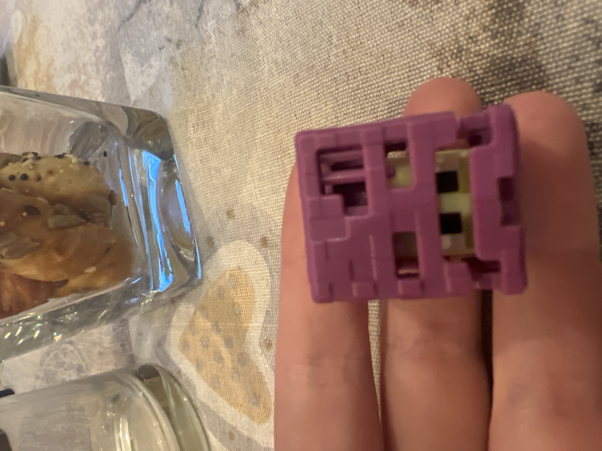 The image captures a close-up, indoor scene featuring a person's hand holding a small, distinct object. The main subjects are a hand and a purple, blocky object held within its fingers, occupying the right side of the frame. On the left, two clear glass or plastic containers are visible. One container, in the upper-left, holds several golden-brown baked goods, possibly crackers or small pastries, some with visible seeds (like poppy or sesame seeds) on their surface. A second, blurry clear container is partially seen in the bottom-left corner.
The purple object held by the hand has a geometric, pixelated design, suggesting it might be a toy or a novelty item. It features various cut-outs and internal structures, revealing hints of light yellow/green and black components inside. The activity taking place is simply a hand holding this object, possibly examining it or preparing to interact with it further.
The setting appears to be a casual indoor environment, likely a table or countertop, covered with a textured fabric. The fabric has a light beige or off-white color with a subtle, abstract pattern of darker, speckled shapes, resembling a tablecloth or placemat. The lighting suggests an ambient indoor light source, but the time of day is not discernible. No specific location cues are visible in the image to confirm the Plovdiv, Bulgaria context. There is no visible text in the image.