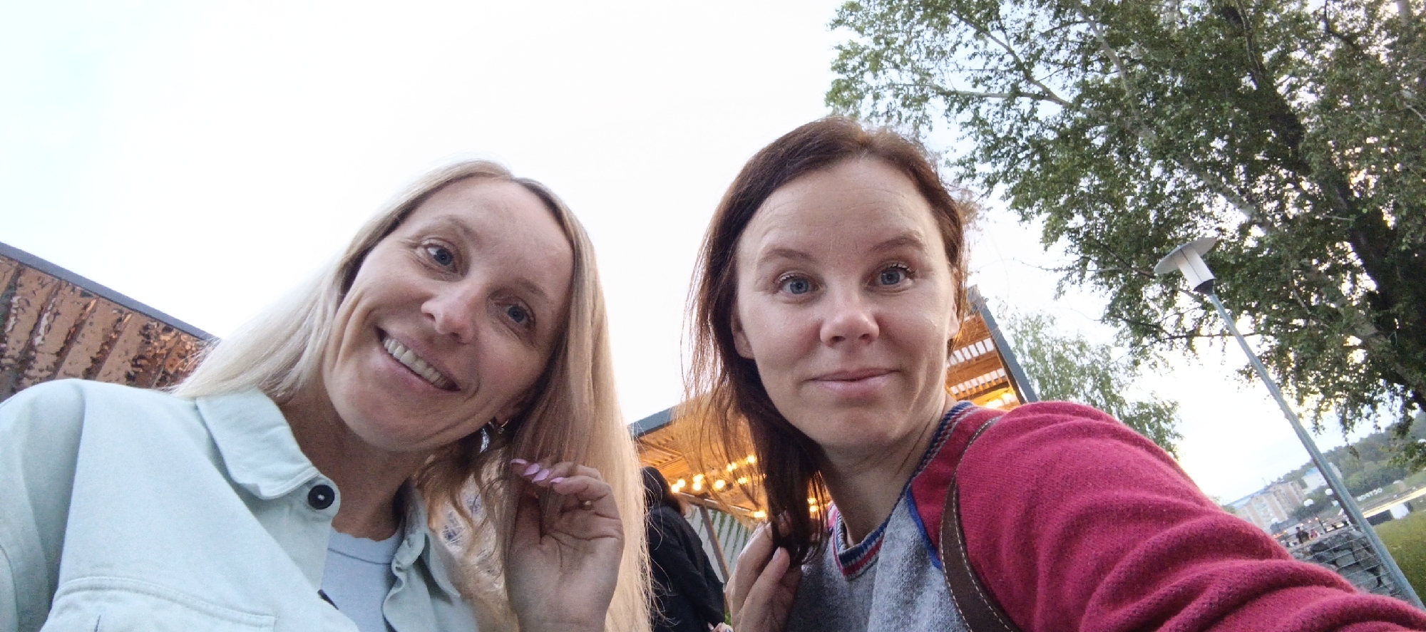 Two women are captured in a close-up, low-angle selfie. The woman on the left has long, blonde hair and is wearing a light blue jacket. She is smiling and looking at the camera. The woman on the right has brown hair and is wearing a pink and gray sweater. She is also looking at the camera with a neutral expression. In the background, there are trees, a street lamp, and buildings that suggest an urban setting. The scene appears to be outdoors, possibly in a park or public area. The presence of streetlights and buildings indicates it is likely within a city. The overall lighting suggests it is either early morning or late afternoon.