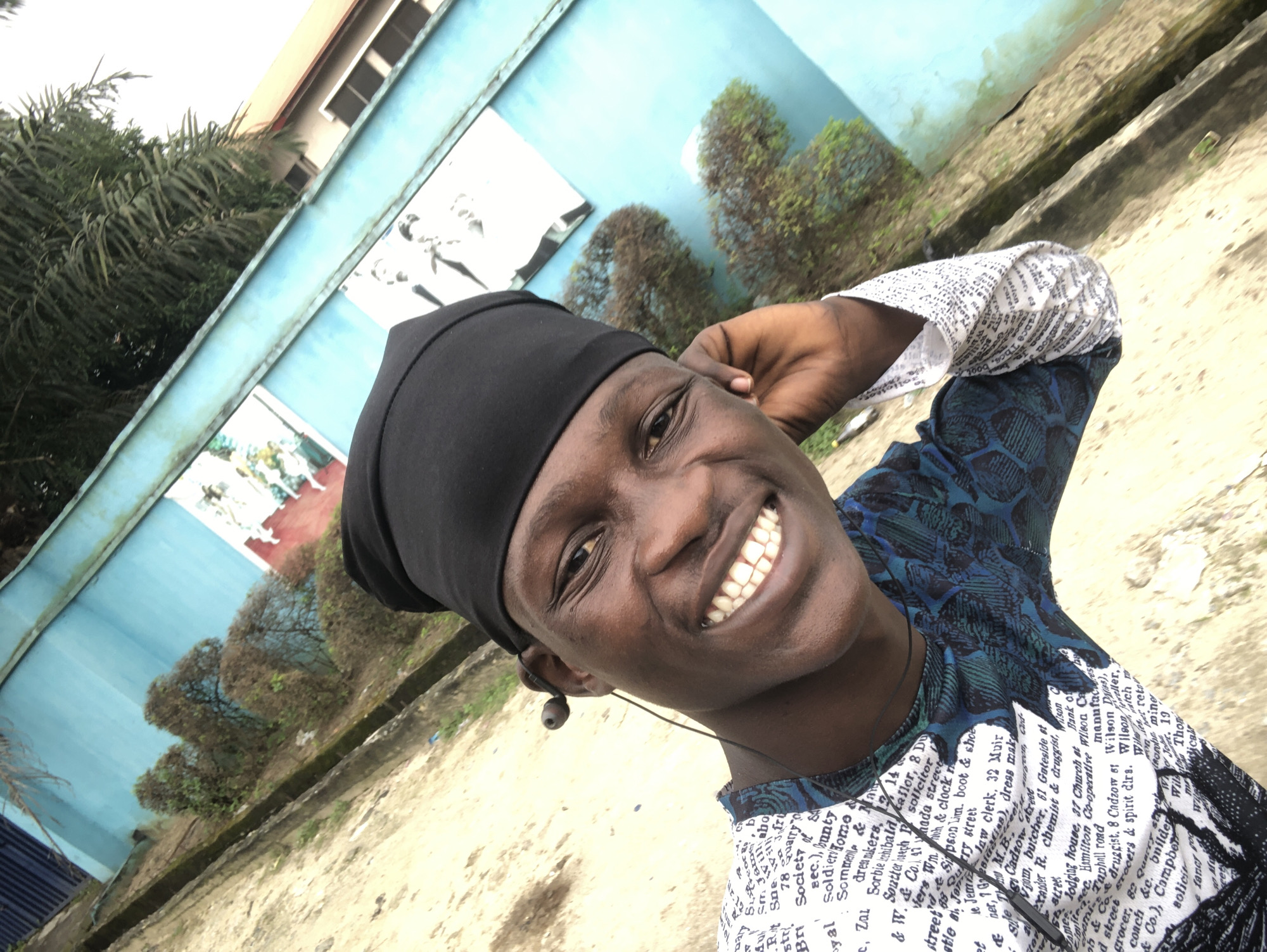 The image is a close-up, selfie-style shot of a young Black male with dark skin, a black head covering, and a black and blue patterned shirt. He is smiling broadly, showing his teeth, and has an earphone in his right ear. His left hand is touching his cheek, and his right hand is not visible. The background is an angled view of a light blue wall with some green foliage and what appears to be a mural or poster on the wall. A section of a tiled roof is visible in the upper right corner. The ground in the foreground is a light brown, textured surface. The lighting is natural, suggesting an outdoor environment. The text on the shirt is predominantly black lettering on a white background, arranged in a repeating pattern of what appears to be names and addresses. The overall impression is a casual, outdoor portrait in an urban setting.