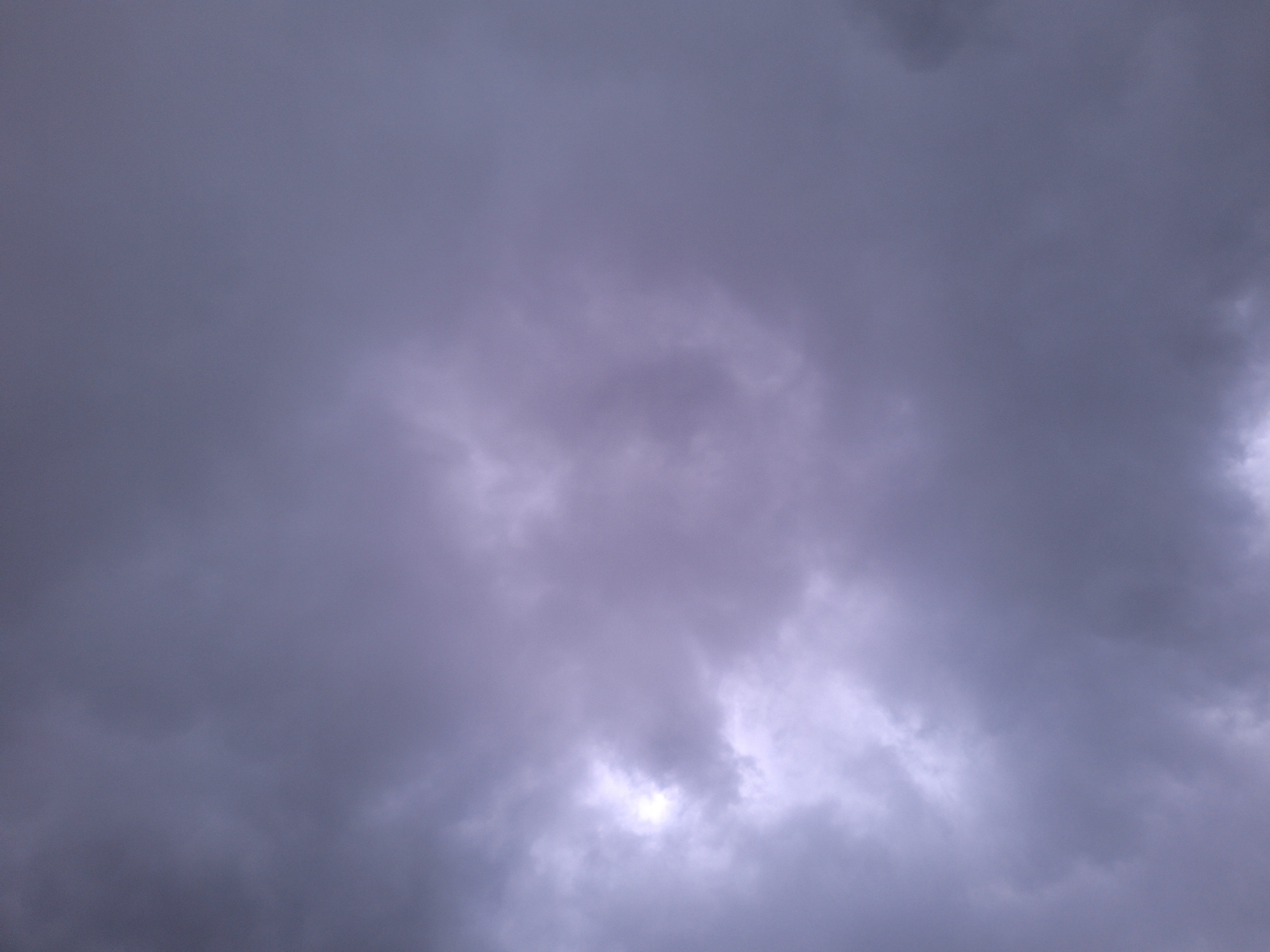 The image shows a sky filled with dense, dark gray clouds. A small area of brighter light is visible through a break in the clouds near the center of the frame, suggesting the sun may be trying to penetrate. The overall impression is of an overcast or stormy sky. There are no people, objects, or discernable landmarks visible in the image.