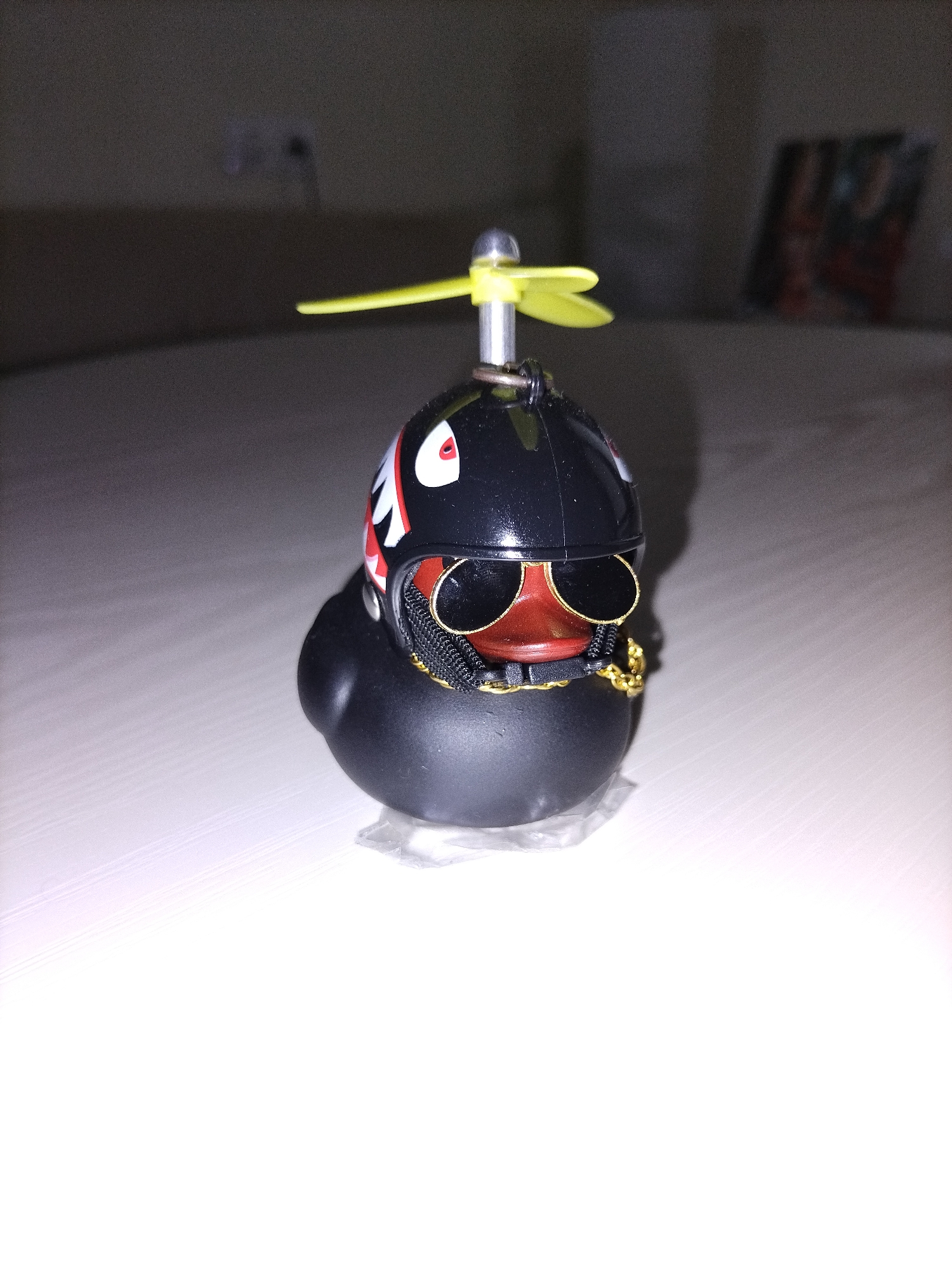 The image features a black rubber duck figurine prominently displayed on a light-colored, reflective surface, likely a table or desk. The duck is accessorized with a glossy black helmet, which has a two-bladed yellow propeller mounted on its top. The helmet's side is decorated with a white and red "shark teeth" motif, reminiscent of military aircraft nose art. The duck also wears black-lensed sunglasses with gold-colored frames, covering its eyes, and a chunky gold chain around its "neck." A small, clear plastic stand supports the duck.
The background is dark and out of focus, suggesting a shallow depth of field and an indoor setting with subdued lighting, possibly evening or night. Vague shapes in the background indicate furniture, such as a light-colored couch or bed, and a blurry rectangular object, possibly a framed picture, is visible on a distant wall to the right. The lighting illuminates the subject clearly, highlighting its details. There is no visible text in the image.