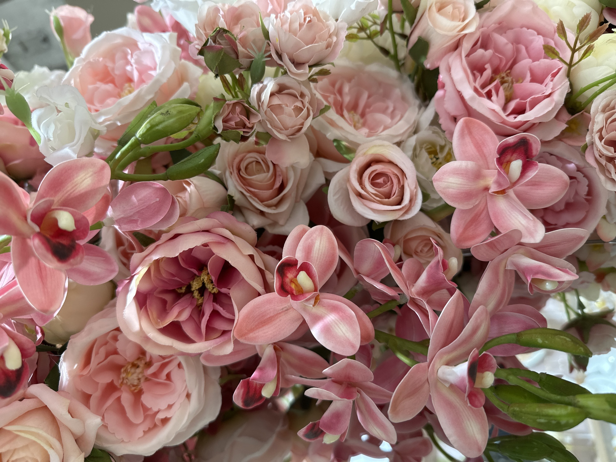 The image presents a vibrant, close-up view of a dense floral arrangement, rich in shades of pink, cream, and white. The composition is dominated by two main types of flowers: roses and orchids.
Numerous roses are visible, ranging from tightly furled buds to fully bloomed flowers with multiple layers of petals. Their hues vary from soft, delicate blush pinks to more saturated rose tones, with some appearing in creamy white. The centers of several open roses reveal fine, yellowish stamens.
Interspersed throughout the arrangement are elegant pink orchids. These orchids display characteristic petaled structures, with many featuring a distinctive darker pink or reddish-brown spot and a creamy-white core on their labellum (lip). Some orchids are fully open, showcasing their intricate forms, while others are still in bud form, appearing as green, elongated capsules on their stems.
The flowers are packed closely together, creating a sense of abundance and lushness. The soft color palette, primarily pastels, gives the arrangement a delicate and sophisticated aesthetic. A subtle, blurred reflection of the flowers is visible on the surface beneath them, suggesting the arrangement rests on a smooth, possibly polished or mirrored, surface indoors. There are no people, discernible text, or specific outdoor location cues present in the image.
