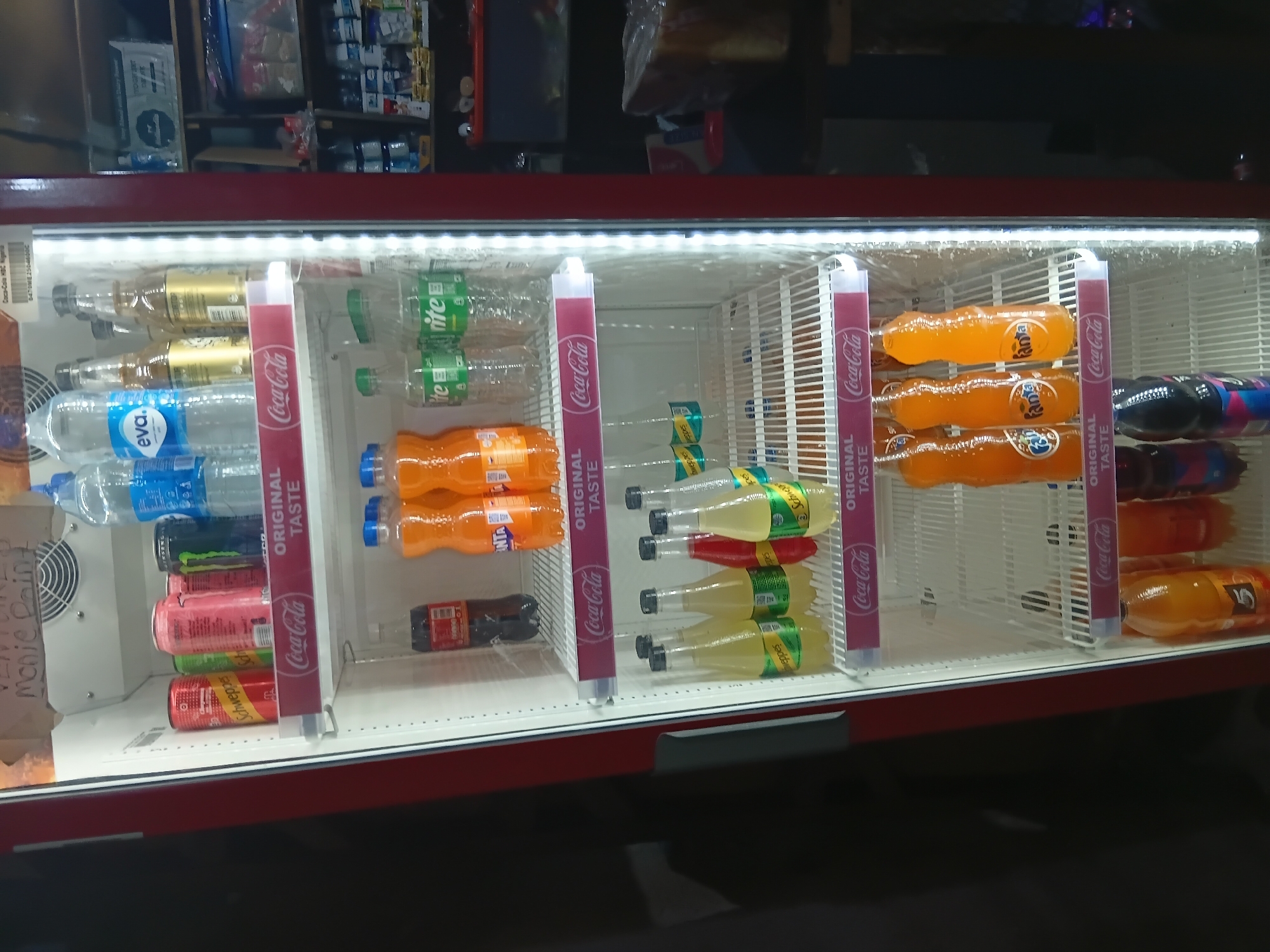 The image depicts a commercial refrigerator filled with various beverages. Located within a store, likely a mini-mart or a small supermarket, this refrigerator is positioned in an aisle. On the top shelf, several bottles of water and cans of Schweppes and Coca-Cola are visible. The middle shelf contains bottles of Fanta, Coca-Cola, and Schweppes. The bottom shelf showcases more bottles of Fanta and other assorted soft drinks. The overall environment suggests a retail setting, possibly a local business in Jos, Nigeria, where such refrigerators are common for displaying cold drinks. The lighting is consistent with an indoor retail environment, with the refrigerator's internal lights illuminating the products. There are no people visible in the frame.