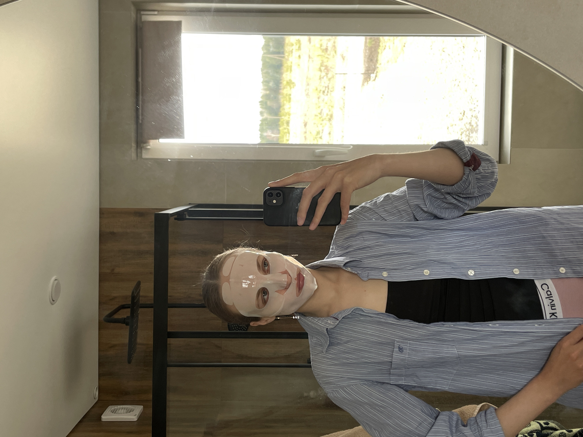 A person, likely female, is taking a mirror selfie indoors, in what appears to be a bathroom or dressing area. They are wearing a translucent sheet face mask, covering their face from forehead to chin, with cutouts for the eyes, nose, and mouth. Their hair is pulled back. The subject is dressed in a light blue and white striped long-sleeved button-down shirt, which is partially unbuttoned to reveal a black bralette or sports bra. A pink waistband, possibly of underwear or shorts, is visible, with the text "Calvin Klein" printed on it. The person holds a black smartphone, featuring multiple cameras, in their right hand to capture the reflection.
The background includes a large window through which bright natural light streams, reflecting green foliage and trees, indicating it is daytime. The room features light-colored walls, dark wooden paneling, and a dark metal frame structure, possibly a shower enclosure or towel rack. A white circular switch or button is present on a white wall to the left. A white square vent or drain cover is seen near the bottom edge against the wooden floor or wall. The setting is in Подшивалово, Russia.