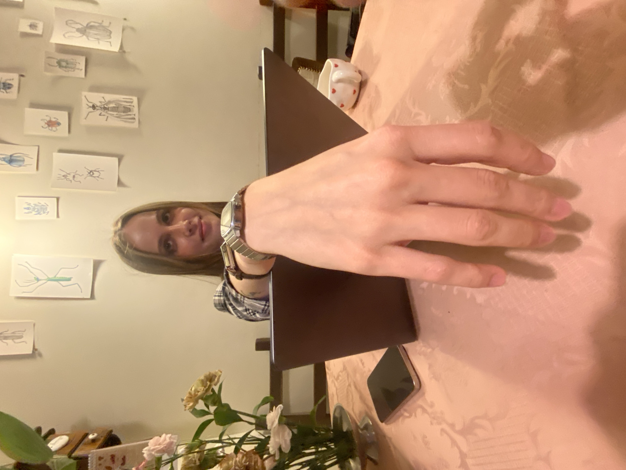 The image captures an indoor scene, likely a home in Moscow, Russia, centered around a young woman seated at a table. The perspective is tilted, showing the woman from a slightly overhead and side angle. She has light hair and is wearing a plaid shirt. Her face is partially visible as she looks towards the camera with a neutral to slight smile. A metallic wristwatch is visible on her left wrist.
Her left hand extends across the foreground, positioned over a dark-colored laptop that is open on the table. To the right of the laptop, a smartphone lies face down on the table. The table is covered with a light-colored, ornate patterned tablecloth. A white mug with small red heart decals is situated behind the laptop.
In the lower left corner, a bouquet of light-colored flowers, possibly carnations or similar, stands in a metallic vase. The background wall is light-colored and decorated with several framed artworks, depicting detailed line drawings of various insects such as beetles, a fly, and a praying mantis, arranged in a gallery style. The lighting in the room appears warm, suggesting an evening or indoor setting with artificial light. No visible text is clearly discernible in the image.