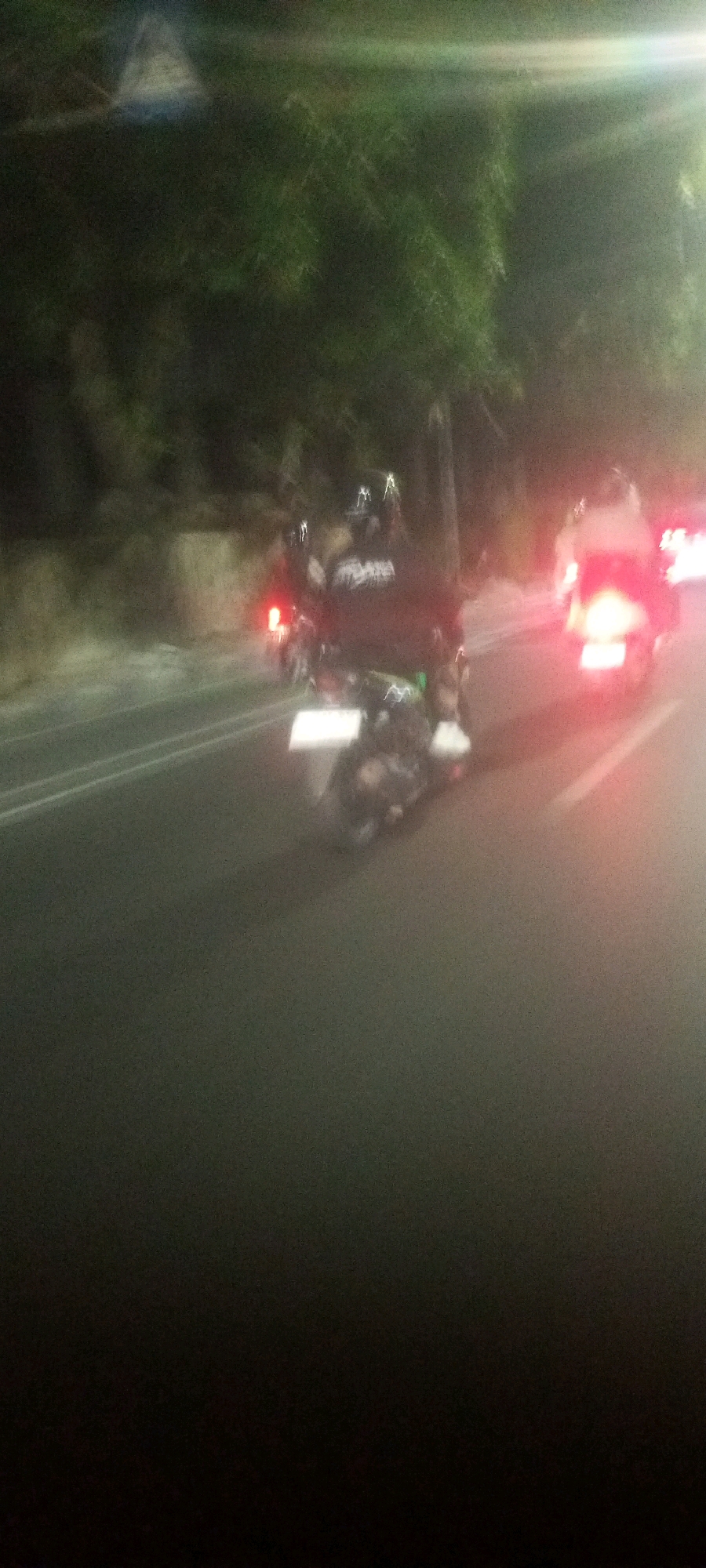 The image depicts a dimly lit, slightly blurred scene of motorcycles on a road, likely at night, given the artificial lighting and low visibility. Two motorcycles are prominently visible in the foreground, with at least two individuals on the lead motorcycle, one appearing to be driving and the other as a passenger. The lead motorcycle has a white license plate visible at the rear. Red taillights are illuminated on both motorcycles. In the background, there is greenery and what appears to be a stone wall or embankment. Further down the road, additional red lights suggest more vehicles are present. The overall impression is of movement on a road in an urban or semi-urban setting, possibly in Indonesia, given the provided location context. There's a bright light source, possibly a streetlight or a vehicle headlight, creating a glare in the upper right quadrant of the image.