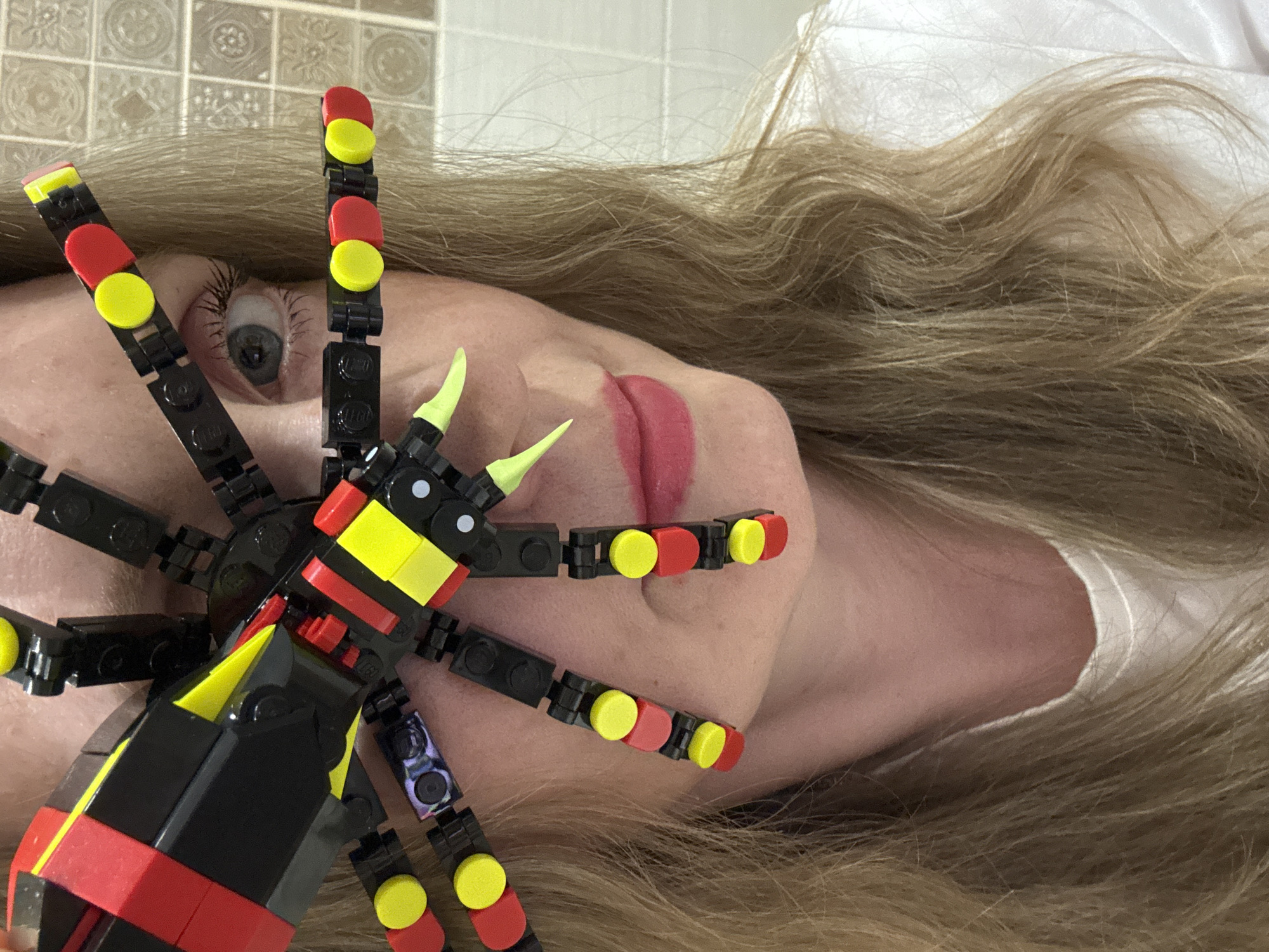 The image depicts a fair-skinned person with blonde hair lying down with a large, colorful LEGO spider positioned on their face. The spider's body is primarily black and red with yellow accents, and it has eight black segmented legs with alternating red and yellow LEGO studs at the end. Two neon yellow "fangs" protrude from the spider's head. The person's face is turned towards the camera; one eye is visible above the spider, and their lips are partially visible. The background shows tiled walls. There is no indication of this being located in Chaikovsky, Russia.