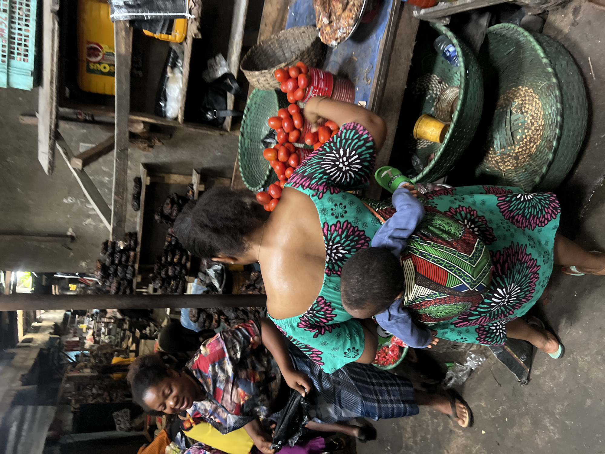 In the city of Nkpor, Nigeria, this photograph captures a vibrant market scene. A woman, facing away from the camera, wears a striking green dress adorned with a floral pattern in pink and white. She is carrying a young child on her back, secured by a colorful fabric wrap featuring geometric designs in red, green, white, and yellow. The child, facing forward, is dressed in a blue long-sleeved shirt and light blue pants.
To the left, another woman, viewed from the side and looking downwards, wears a patterned shirt with red, black, and white elements and a blue and white checkered skirt. She holds a dark object in her hands, possibly a bag.
The foreground is dominated by a stall laden with fresh produce. Several cans are filled with ripe red tomatoes, stacked in rows. To the right, a shallow bowl contains what appears to be dried fish. Below the stall, two large, dark green woven baskets are stacked, the lower one with a textured lid. A smaller green woven basket sits above them, containing a yellow container and other items.
The market environment is rustic, with wooden shelves and structures visible in the background. The ground is a rough, concrete surface. The overall impression is one of bustling commerce and daily life in a Nigerian market.