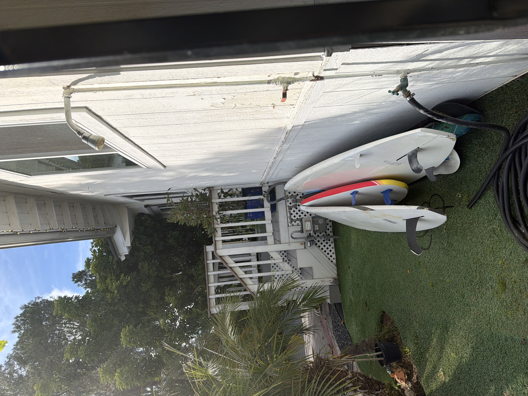 The image depicts a scene on a sunny day, likely in a residential area near the coast. The most prominent feature is a stack of surfboards leaning against the side of a white, one-story building. The surfboards are of various colors and designs. A water hose is connected to an outdoor faucet on the building. The ground is covered with artificial turf. A set of stairs leads up to the side of the building, and lush trees and vegetation are visible in the background, suggesting proximity to a beach or coastal environment. The building's design and the surrounding vegetation hint that the location could be Malibu, United States of America.