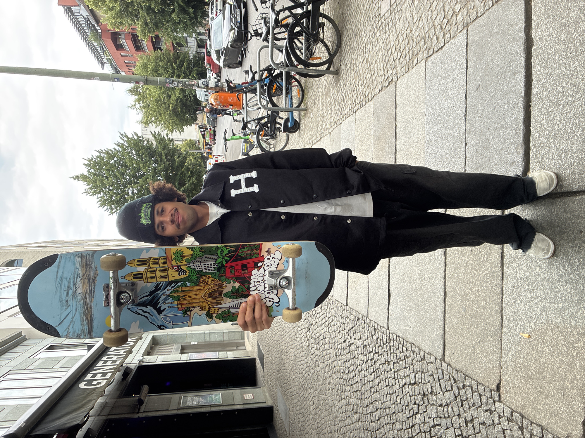 A young man stands on a sidewalk in Berlin, Germany, holding a skateboard. He is wearing a black beanie, a black jacket with a white "H" logo, dark pants, and light-colored shoes. The skateboard has a colorful graphic depicting famous landmarks like the Sagrada Familia, the Golden Gate Bridge, and the Brandenburg Gate. The man is standing in front of a building with a sign that reads "GENERATOR". The sidewalk is paved with cobblestones on the left and large grey stones on the right. In the background, there are trees, buildings, and parked bicycles. The sky is overcast.