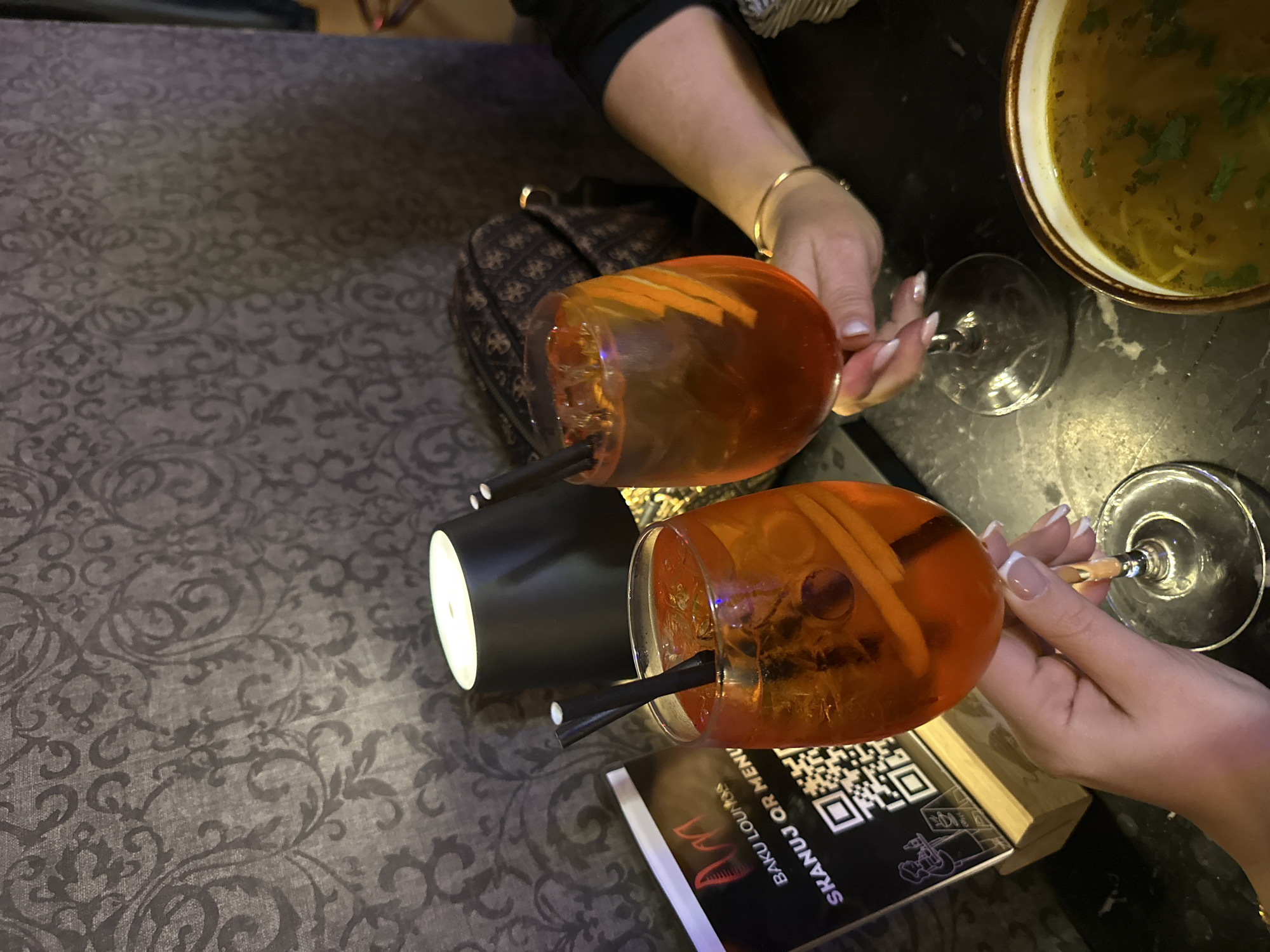 The image shows a high-angle view of a table in what appears to be a restaurant or bar. Two Aperol spritzes are prominently featured, each garnished with orange slices and equipped with a black straw. One drink is held by a hand with neatly manicured nails, while another hand with a gold bracelet holds the other drink. Next to the drinks is a black menu with a QR code and the words "BAKU LOUNGE SKANUJ QR MENU." A white and black cylindrical object is visible beside one of the drinks. In the background, a bowl of soup is positioned on the table. The setting appears to be a dimly lit interior with a patterned tablecloth. Based on the menu visible, it is most likely in Baku Lounge Wrocław, Poland.