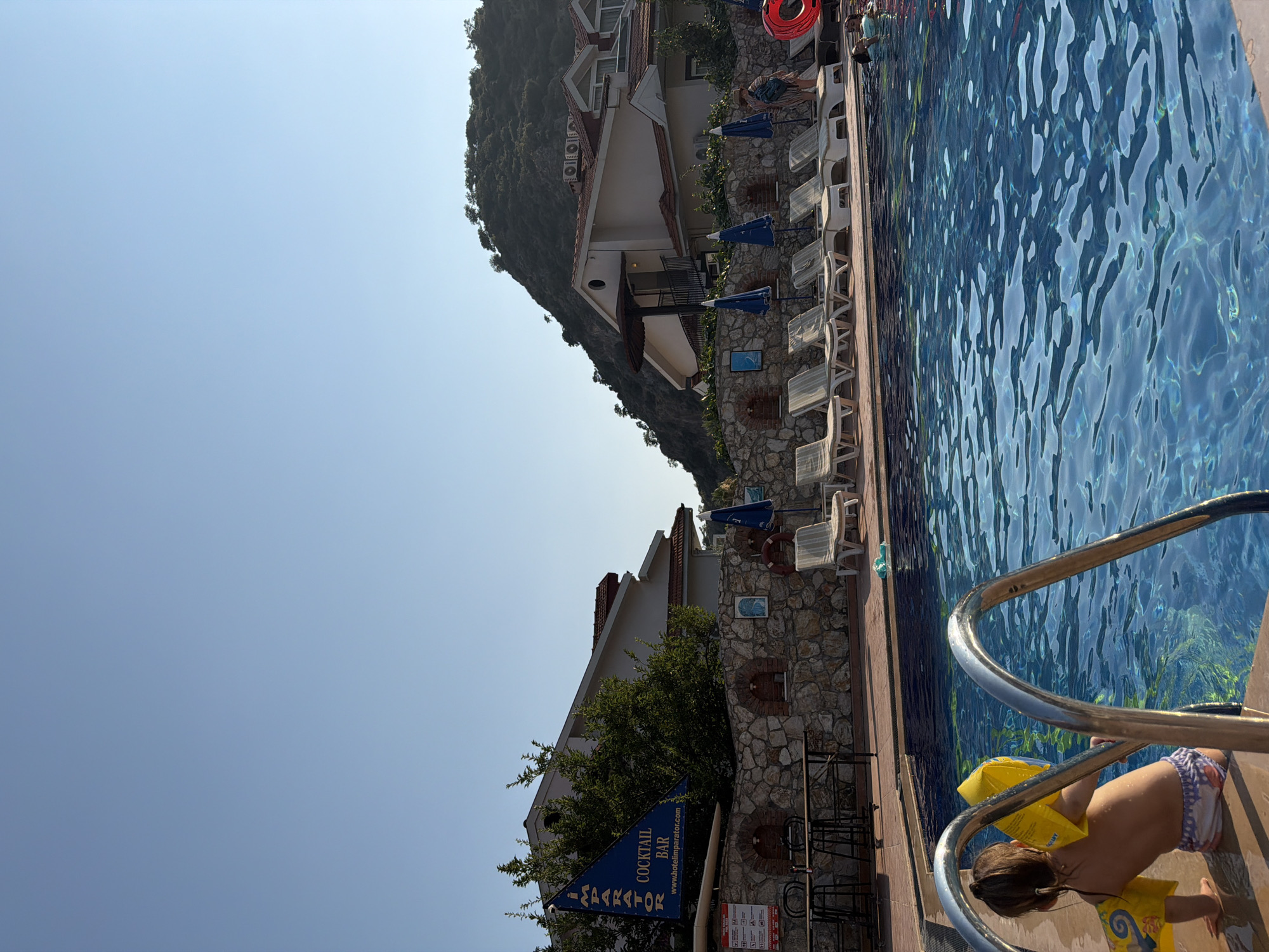 A bright, sunny day is captured at the outdoor swimming pool of the Hotel Imperator in Fethiye, Turkey. The foreground shows a child, wearing yellow armbands and a swimsuit, sitting on the edge of the pool, partially submerged. A metal ladder extends into the clear blue water. In the background, a row of white lounge chairs is set up on the pool deck, some occupied by sunbathers. Beyond the pool area, a stone wall supports a landscaped garden and several buildings, including a multi-story hotel with terracotta roofs, nestled against a verdant hillside. A blue flag with the text "IMPERATOR COCKTAIL BAR" is visible, along with a sign with various icons and information. Umbrellas provide shade near the lounge chairs, and a red inflatable ring is also present. The sky is a clear, pale blue.