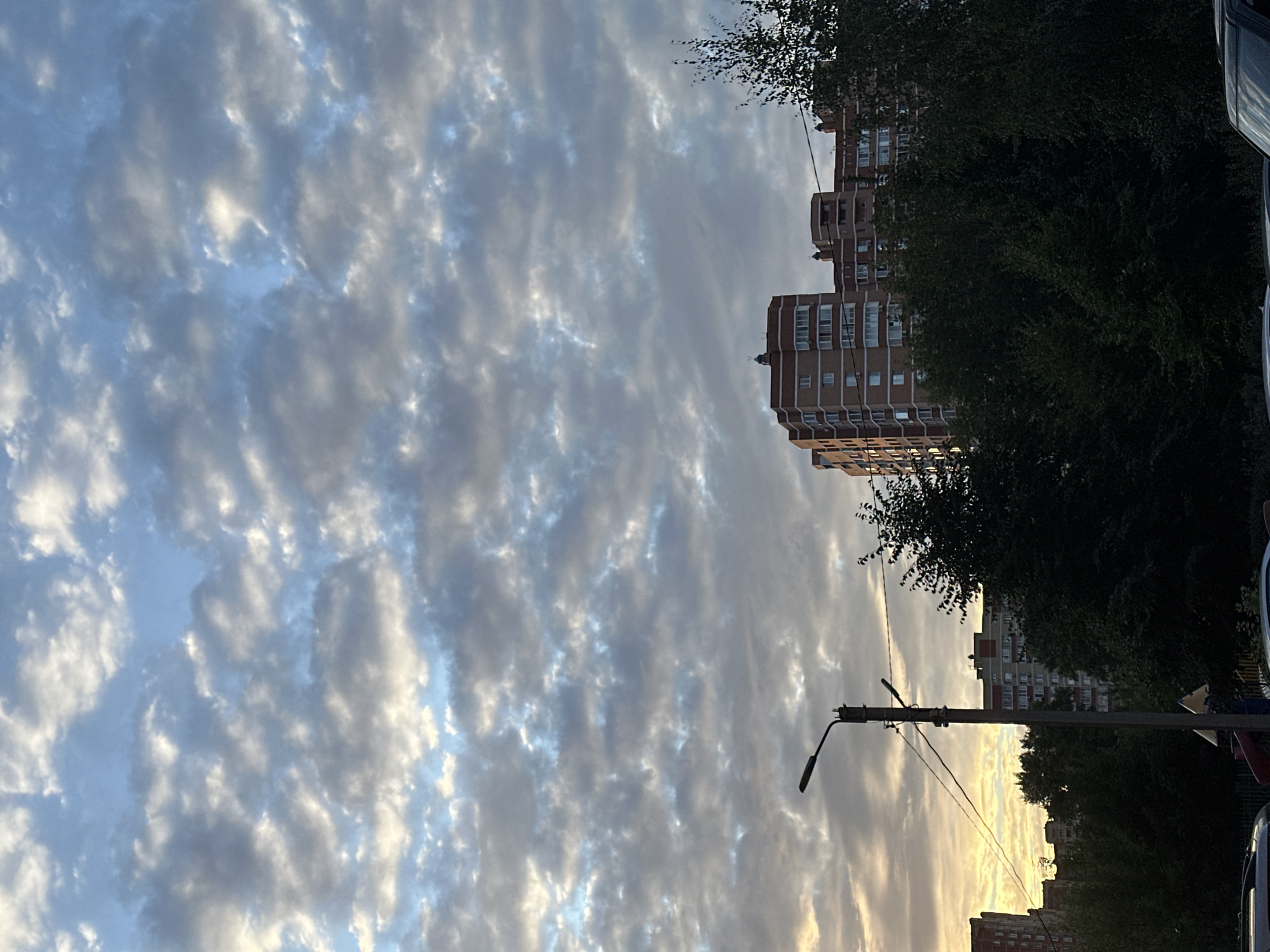 The image depicts a cityscape in Shchyolkovo, Russia, dominated by a dramatic cloudy sky. The sky is filled with numerous cumulus clouds, some appearing bright white and others darker gray, interspersed with patches of blue sky. A warm, golden light, characteristic of sunset or sunrise, is visible on the left side of the frame, illuminating the undersides of the clouds.
In the midground, several buildings are visible. A tall, multi-story brick apartment building stands out with its red-orange facade and numerous windows. Another, lighter-colored apartment building is partially visible to its left. A dark green canopy of trees partially obscures the lower portions of these buildings.
In the foreground, a tall, slender concrete lamppost with two lamps is positioned prominently on the left side of the image. Wires are strung between the lamppost and other unseen points. The base of the lamppost is partially visible, with a small triangular sign and a hint of a playground structure below. The very bottom of the frame shows the hoods of two dark-colored cars. The overall scene is urban, with a focus on the interplay of architecture, nature, and the sky.