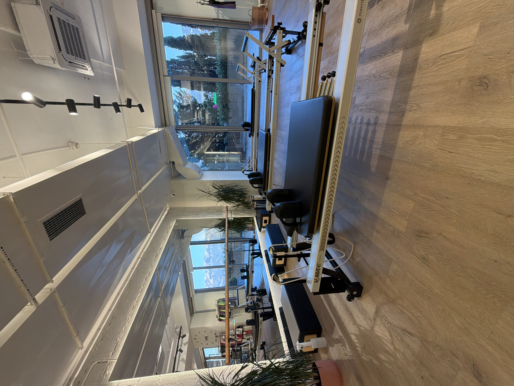 This is a photograph of a Pilates studio located in Boreti, Montenegro. The studio is spacious and modern, with large windows offering a view of snow-capped mountains and trees. The room is filled with several Pilates reformers, the primary equipment for Pilates exercises. These reformers are lined up in rows, some in the foreground and others extending into the background. The flooring is light-colored wood. The lighting appears to be a mix of natural light from the windows and overhead artificial lighting, including track lighting and a ceiling fan or air conditioning unit. There are no people visible in the image. The overall impression is one of a clean, well-equipped, and serene fitness space.